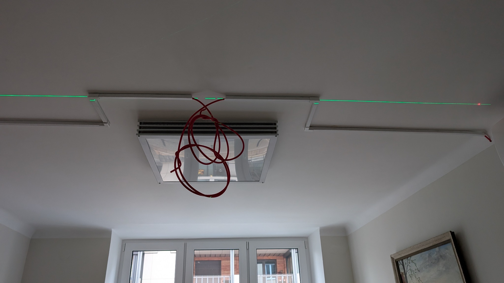 Montaż oświetlenia sufitowego: widoczne profile, lampy w kasetonach i przewody. Użycie lasera do wyznaczania linii. Etap prac instalacyjnych w jasnym wnętrzu.