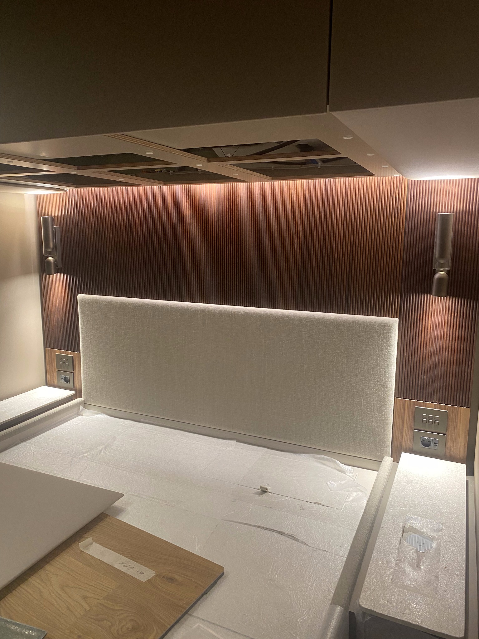 Zamontowane lampy ścienne i gniazdka elektryczne po obu stronach wezgłowia łóżka z drewnianym panelem dekoracyjnym w tle. Prace wykończeniowe w sypialni.