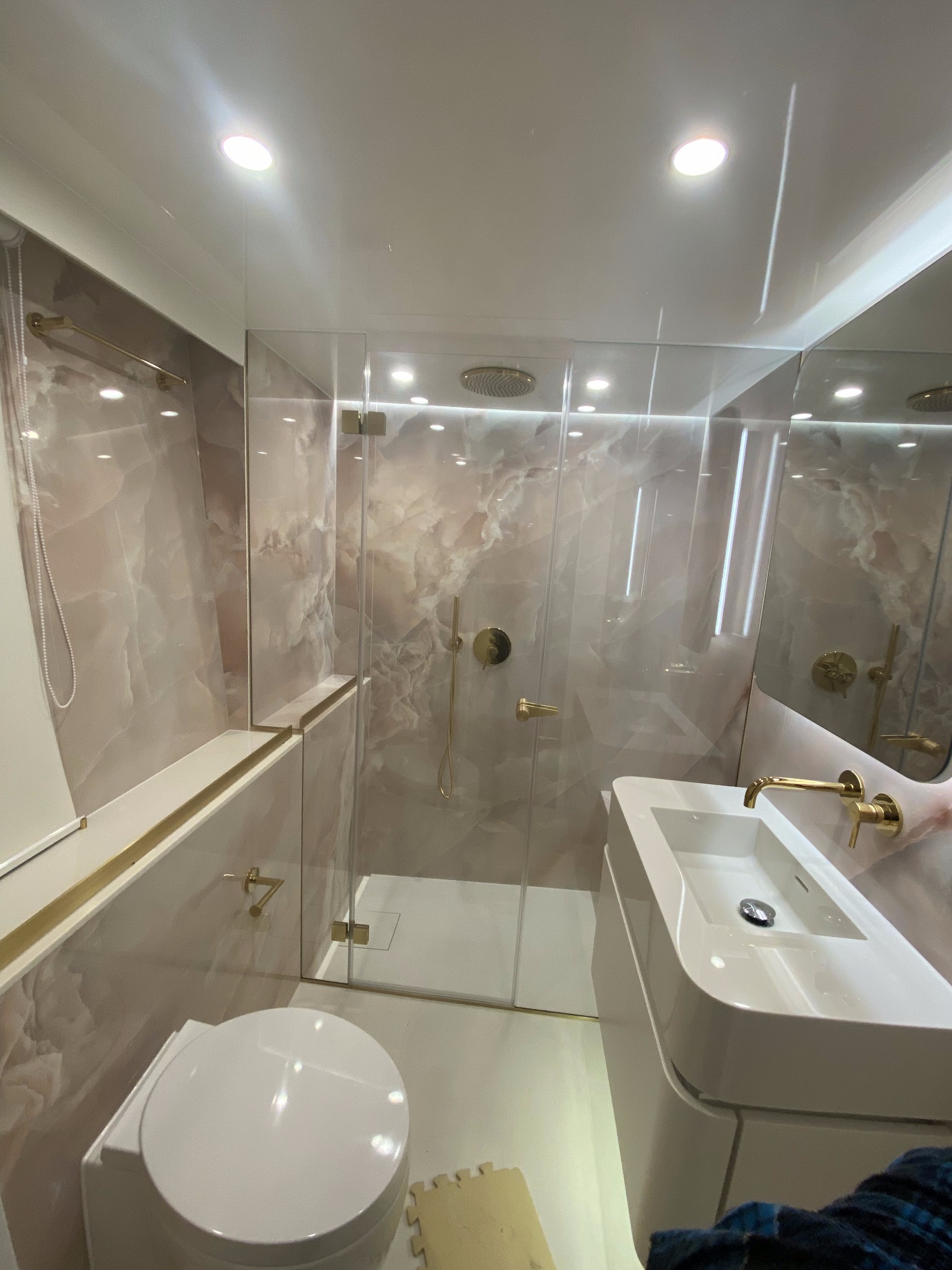 Nowoczesna łazienka po remoncie: marmurowe ściany, szklana kabina prysznicowa z złotą armaturą i oświetleniem punktowym. Biała umywalka z szafką i toaleta.