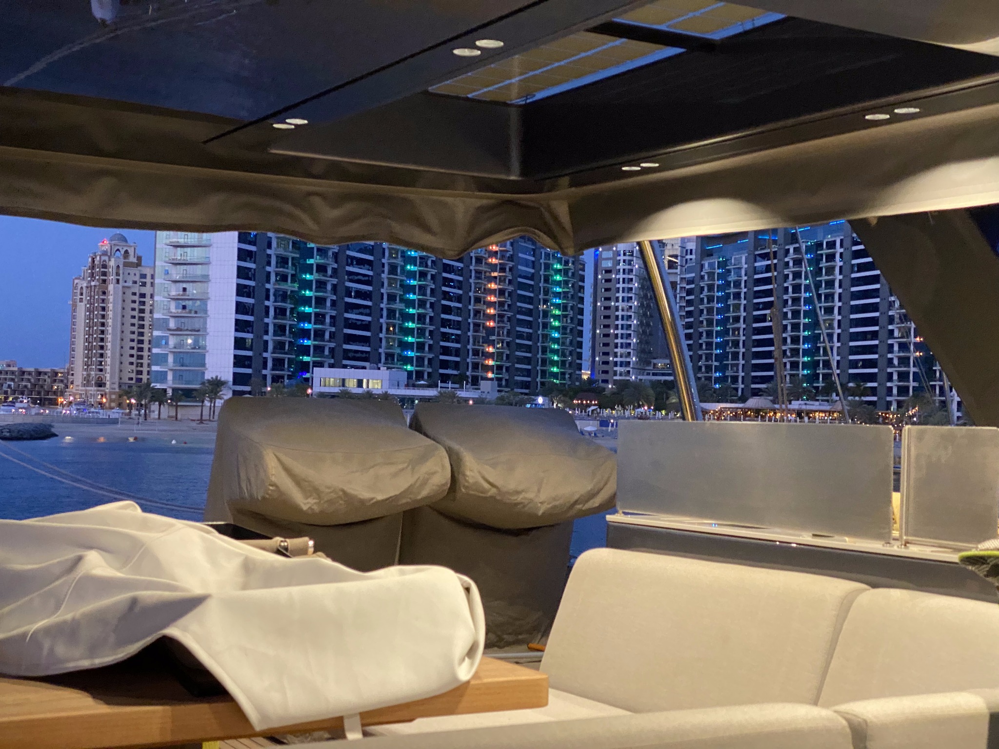 Wnętrze jachtu z widokiem na nocne miasto z podświetlonymi budynkami. Widoczne meble wypoczynkowe i elementy konstrukcyjne jachtu.