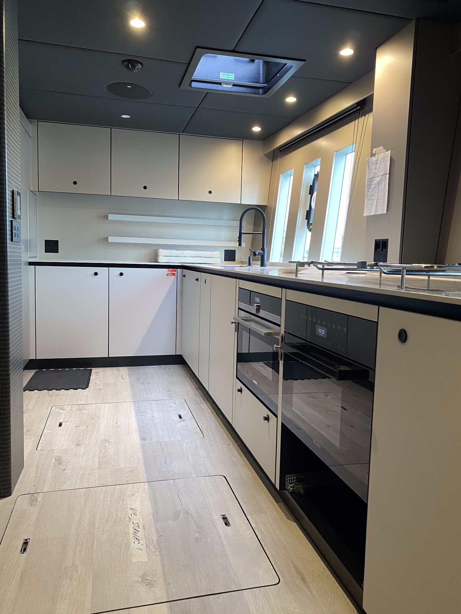 Nowoczesna kuchnia w jachcie z oświetleniem sufitowym, zabudową meblową w odcieniach szarości i czarnymi akcentami armatury oraz sprzętu AGD.