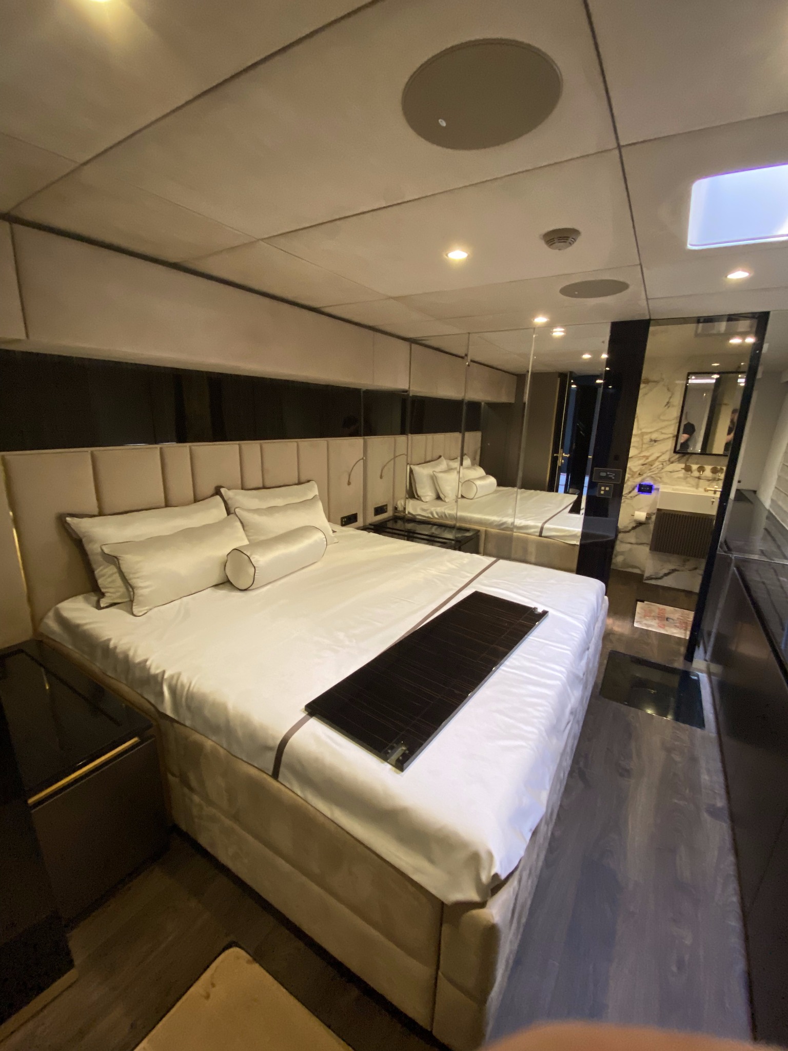 Nowoczesna sypialnia z oświetleniem punktowym w suficie, lustrzanymi ścianami i łazienką w tle. Widoczne gniazdka elektryczne przy łóżku.