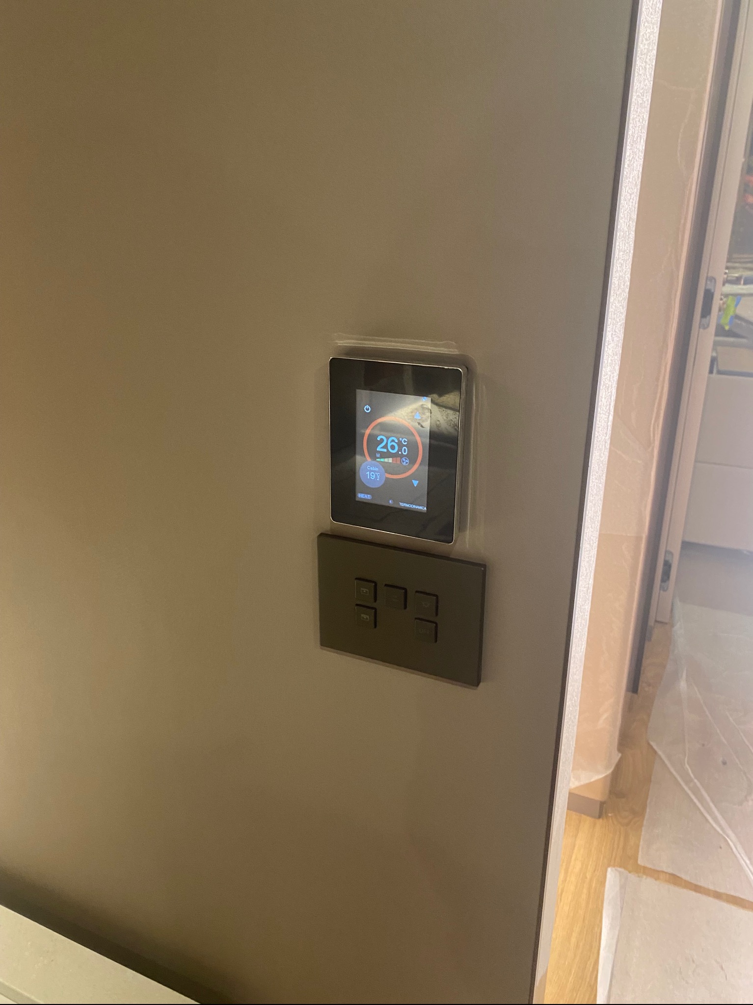 Nowoczesny panel sterowania temperaturą i włącznik światła, zamontowane na szarej ścianie. Widoczny wyświetlacz z temperaturą 26 stopni Celsjusza.