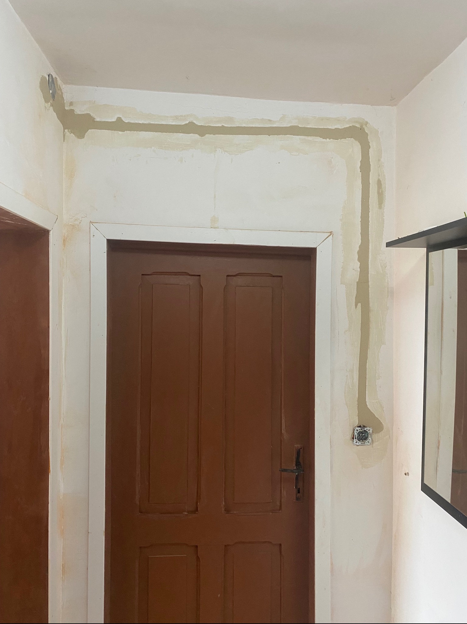 Przygotowanie ściany pod montaż instalacji elektrycznej wzdłuż futryny drzwi i pod sufitem. Widoczne kable i puszka w ścianie, ślady po szpachlowaniu.