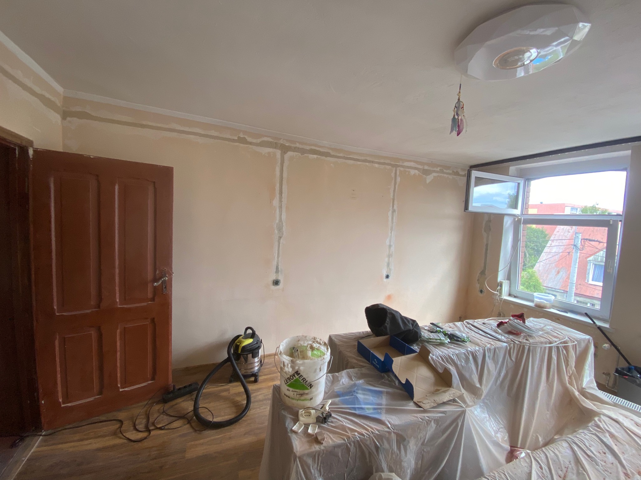 Pokój w trakcie malowania: ściany z widocznymi śladami szpachlowania, zakryte folią meble, otwarte okno, wiadro z farbą Leroy Merlin i odkurzacz. Widoczne kable elektryczne.