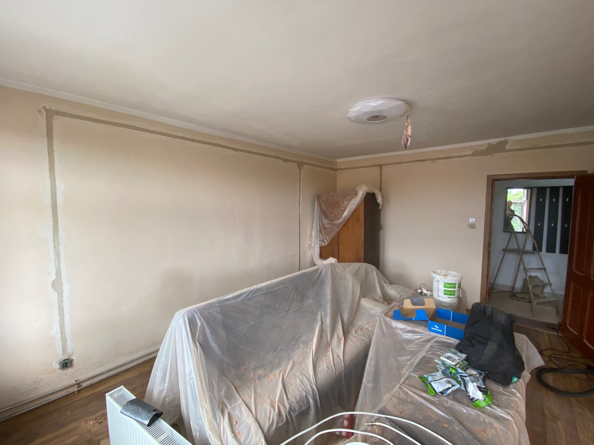 Pokój w trakcie malowania: ściany zagruntowane, meble zakryte folią ochronną, widoczne puszki z farbą i drabina w tle przy otwartych drzwiach.