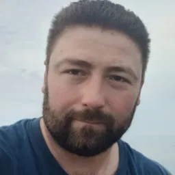 Portret brodatego mężczyzny w niebieskiej koszulce na tle nieba. Ujęcie z bliska, skupione na twarzy i wyrazie oczu. Delikatne oświetlenie.