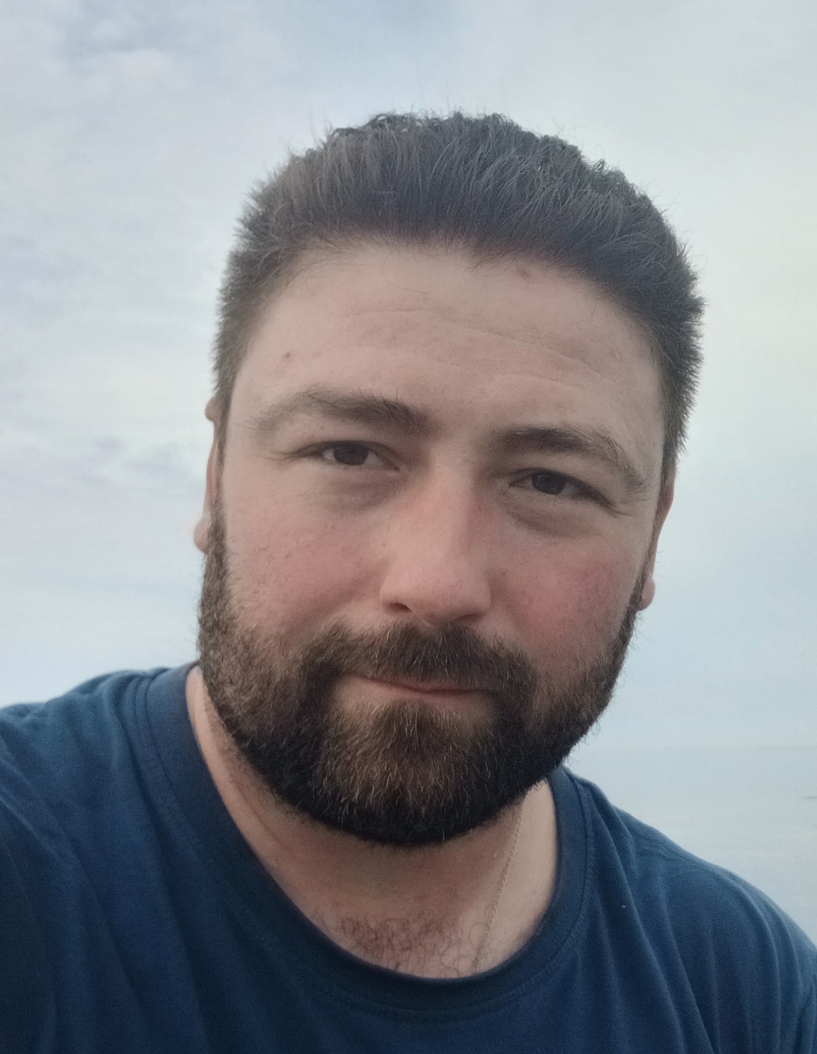 Portret brodatego mężczyzny w niebieskiej koszulce na tle nieba. Ujęcie z bliska, skupione na twarzy i wyrazie oczu. Delikatne oświetlenie.