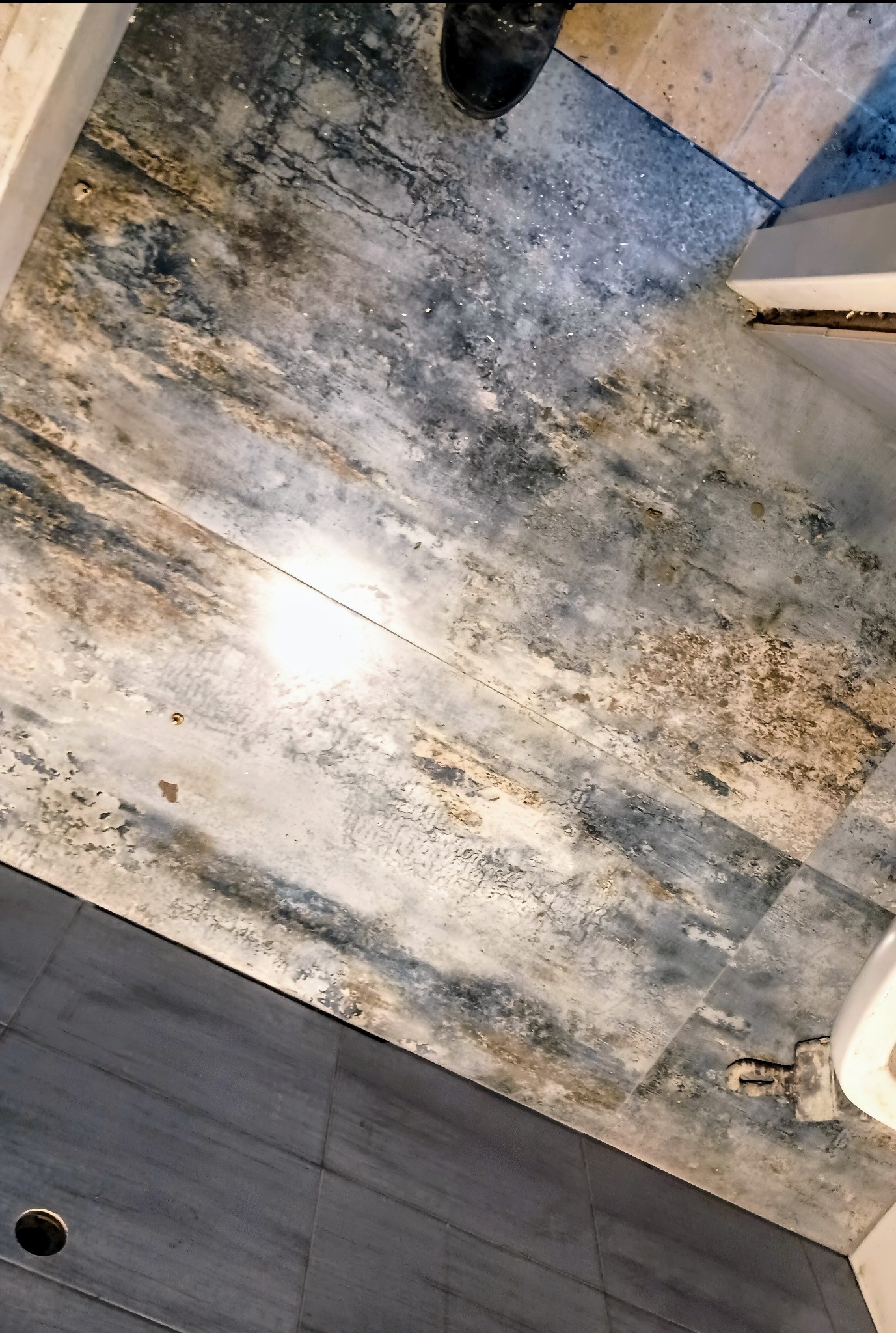 Podłoga w łazience z widocznymi śladami wilgoci i uszkodzeń. Fragment toalety i buta w kadrze. Widoczne różnice w kolorze i fakturze płytek.