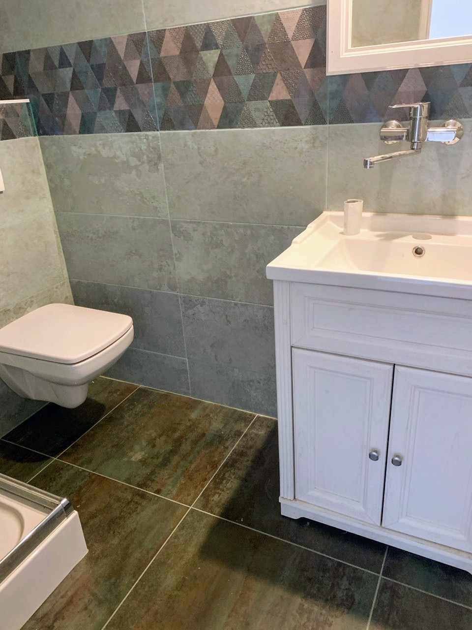 Wykończona łazienka z wiszącą toaletą, umywalką z szafką i geometrycznymi płytkami. Brązowe płytki podłogowe imitujące drewno.