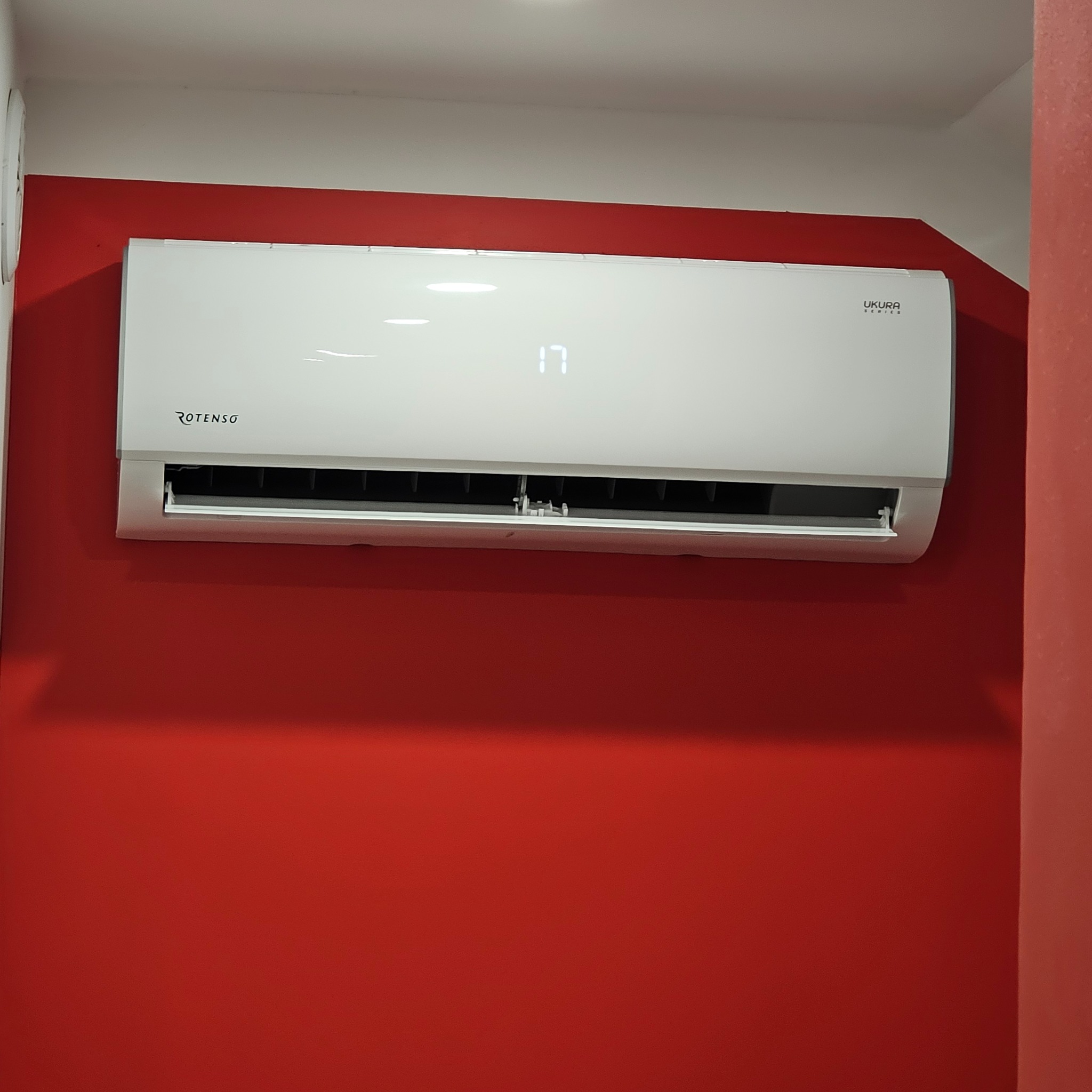Biały klimatyzator Rotenso Ukura Series zamontowany na czerwonej ścianie. Na wyświetlaczu widoczna temperatura 17 stopni.