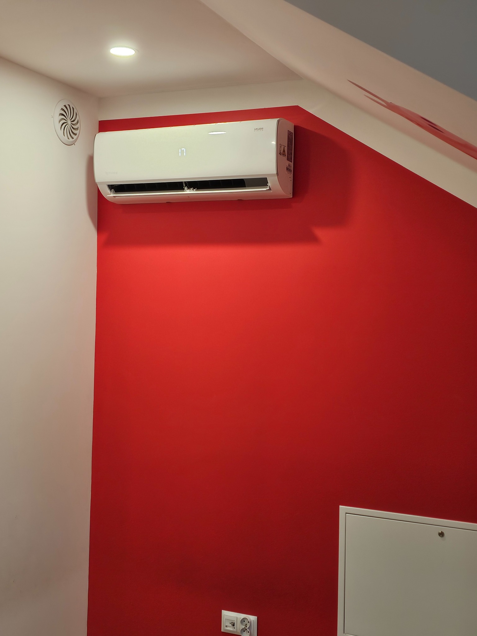 Biała klimatyzacja split na czerwonej ścianie w pokoju. Widoczny wyświetlacz temperatury, kratka wentylacyjna i gniazdko elektryczne. Montaż w rogu pomieszczenia.