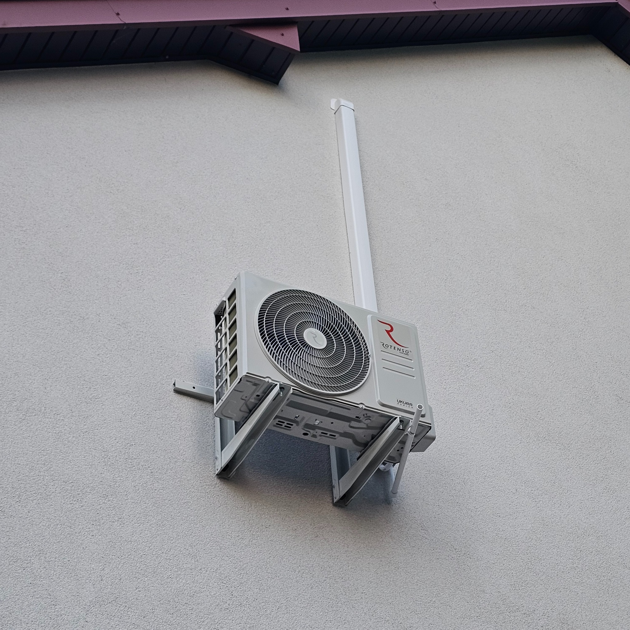 Zamontowana jednostka zewnętrzna klimatyzacji Rotenso na szarej, teksturowanej ścianie budynku. Widoczny biały przewód i fragment dachu. Ujęcie z dołu.