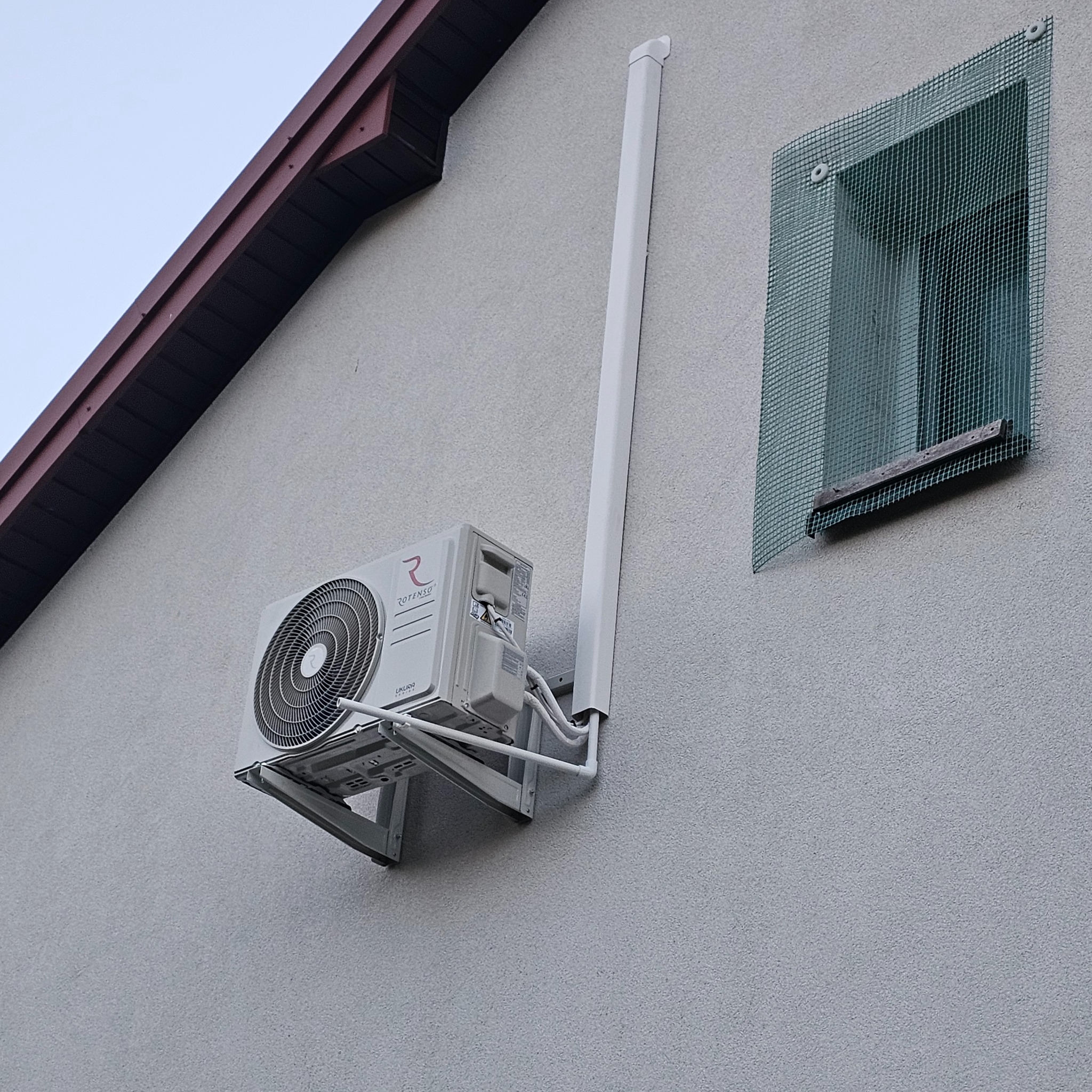Biały klimatyzator Rotenso zamontowany na ścianie domu z szarą elewacją, widoczna rura odprowadzająca. Obok okno z siatką ochronną.