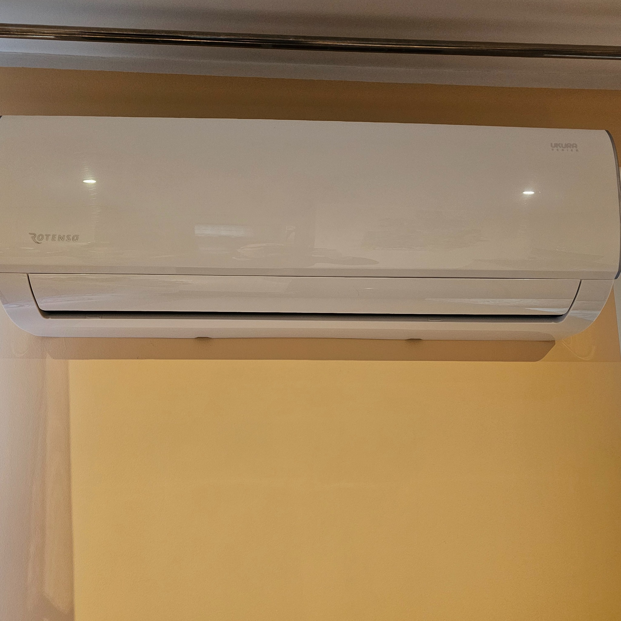 Biała klimatyzacja Rotenso Ukura Series zamontowana na beżowej ścianie w pomieszczeniu. Widoczne logo producenta.