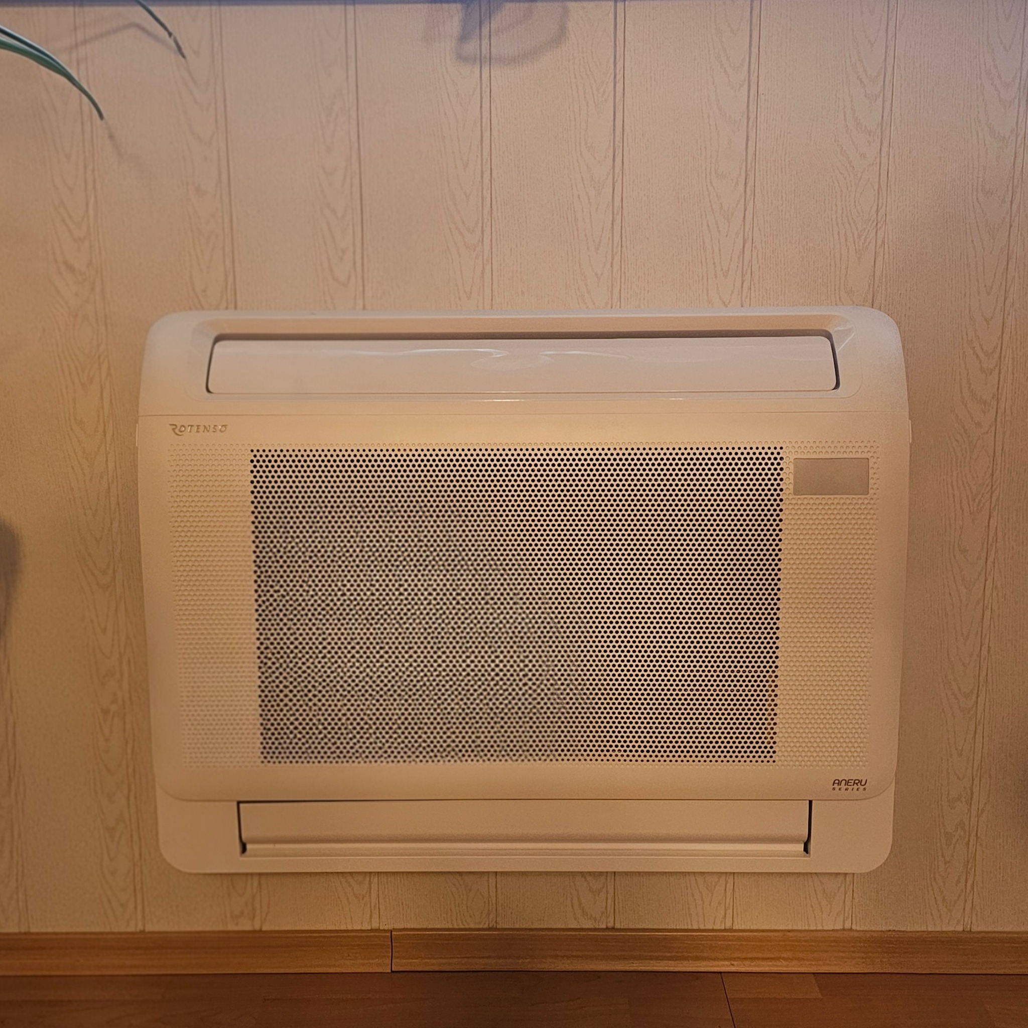 Ścienny klimatyzator Rotenso w jasnym odcieniu beżu, zamontowany na panelowej ścianie z drewnopodobnym wzorem, widok z przodu, dolne ujęcie.