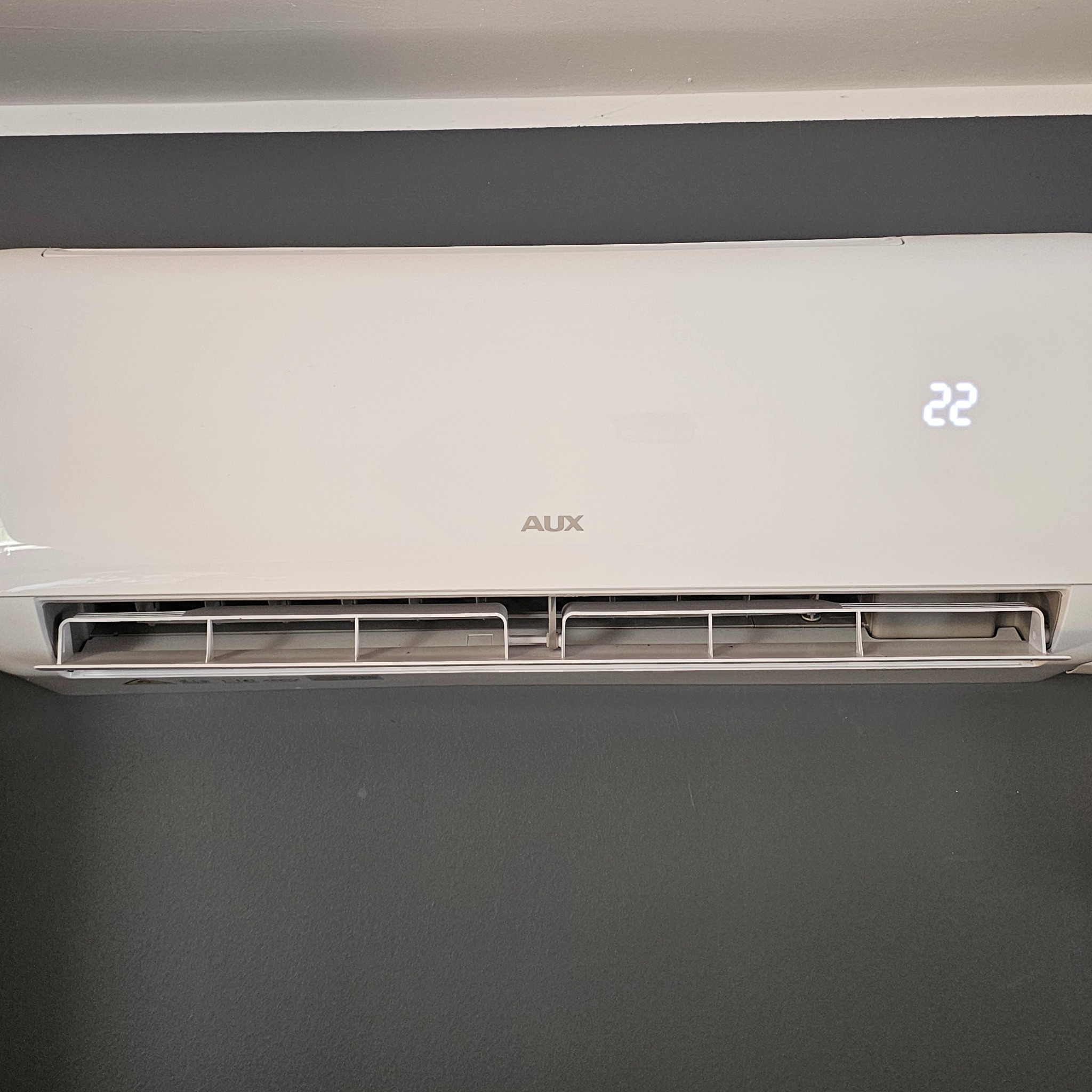 Biała klimatyzacja AUX na szarej ścianie, wyświetlająca temperaturę 22 stopnie. Widoczne szczeliny wentylacyjne i logo producenta.