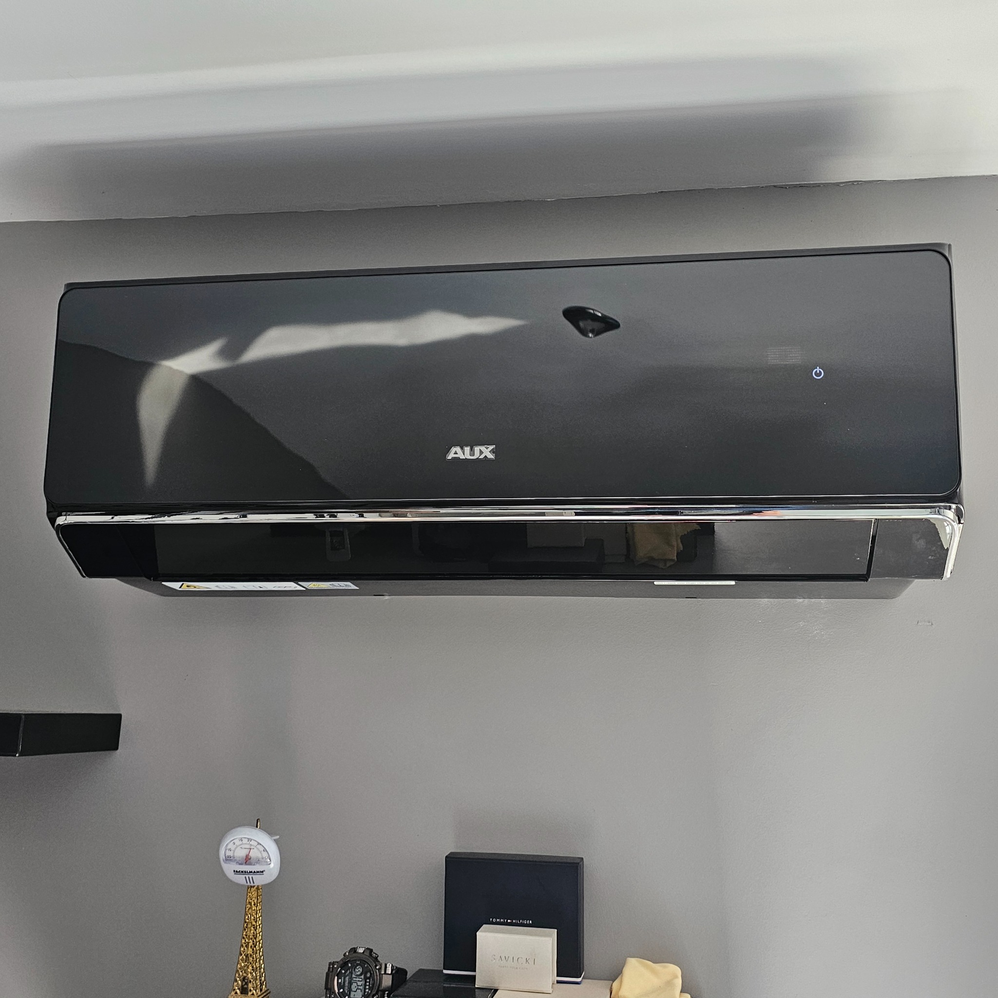 Czarny klimatyzator AUX zamontowany na szarej ścianie w pokoju. Pod nim półka z dekoracjami. Widoczne odbicia światła na obudowie.