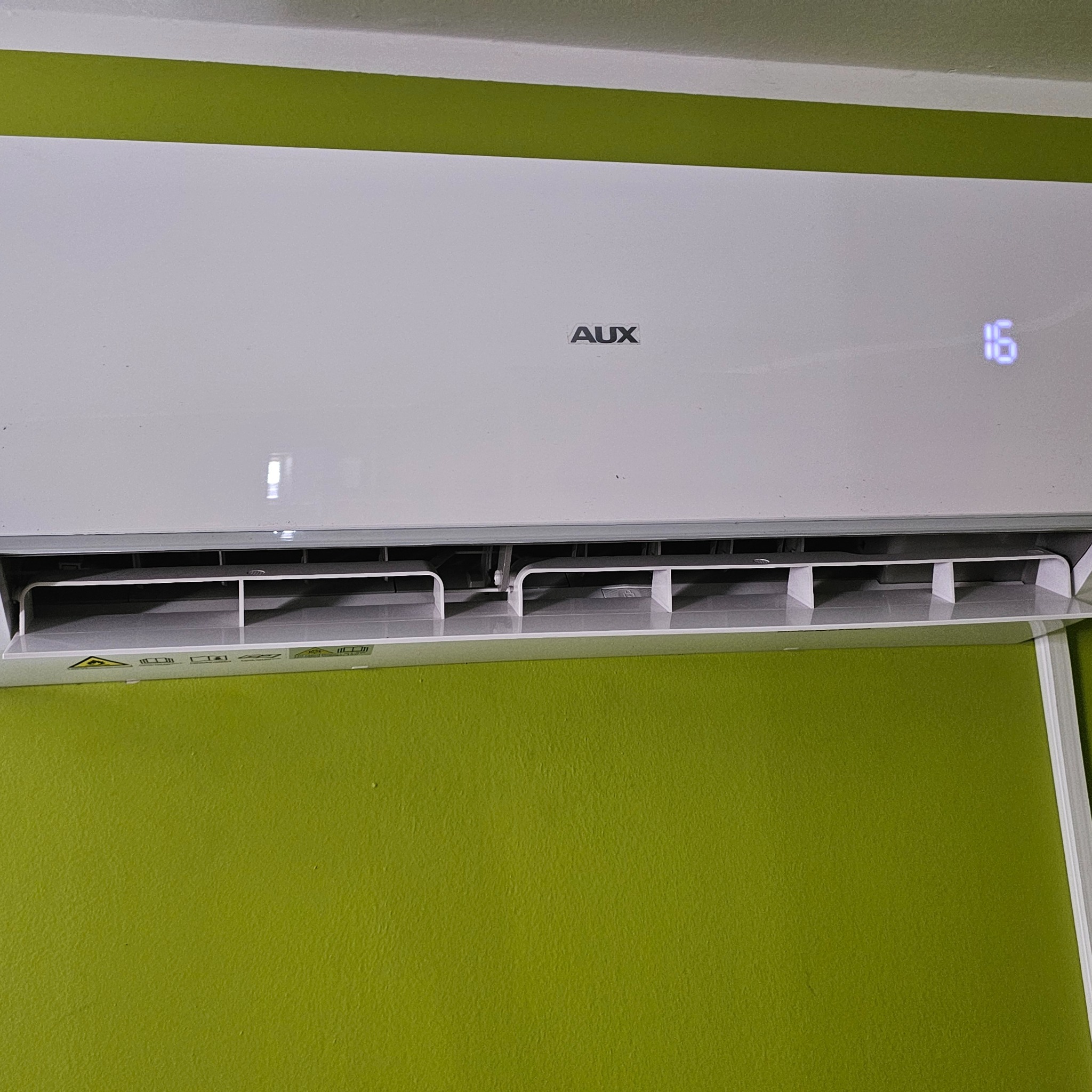 Biała klimatyzacja AUX na zielonej ścianie z wyświetlaczem temperatury ustawionym na 16 stopni. Widoczne żaluzje nawiewu.