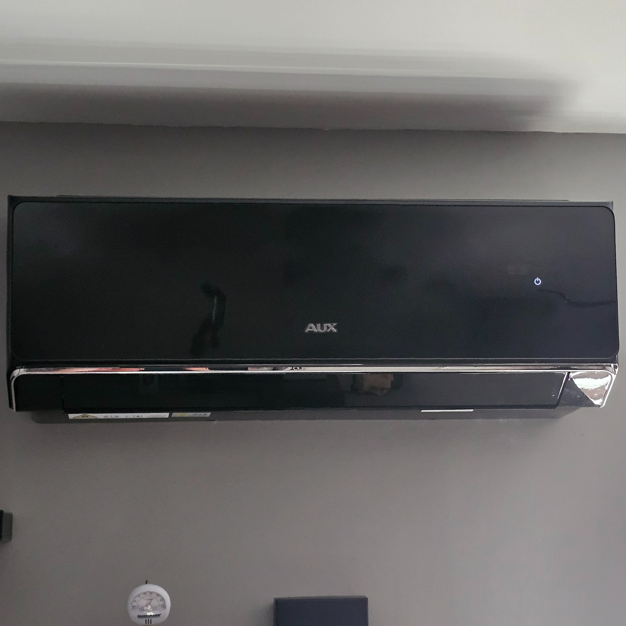 Czarna klimatyzacja AUX zamontowana na szarej ścianie, widoczny termometr i fragment pudełka. Górny kadr.