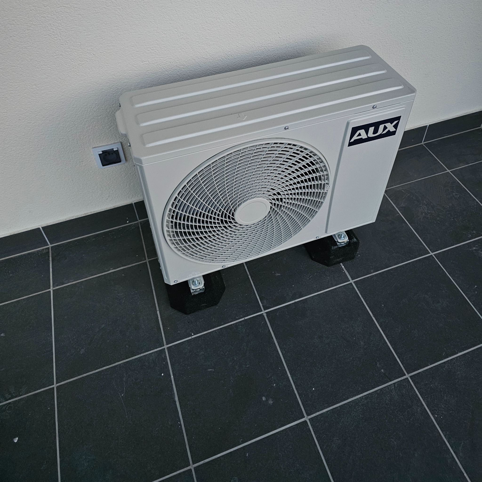Zewnętrzna jednostka klimatyzacji AUX zamontowana na czarnych podkładkach, widoczny fragment ściany z gniazdkiem elektrycznym i ciemna, kwadratowa podłoga.