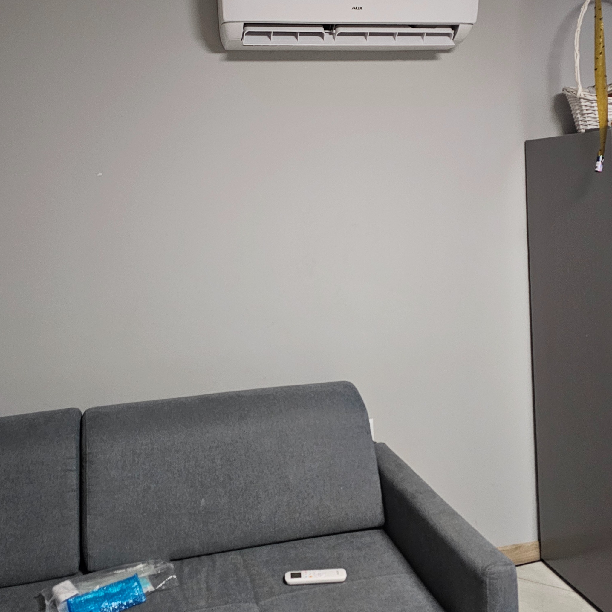 Biały klimatyzator AUX zamontowany na szarej ścianie nad szarą kanapą z pilotem. W tle szafka i koszyk wiklinowy.