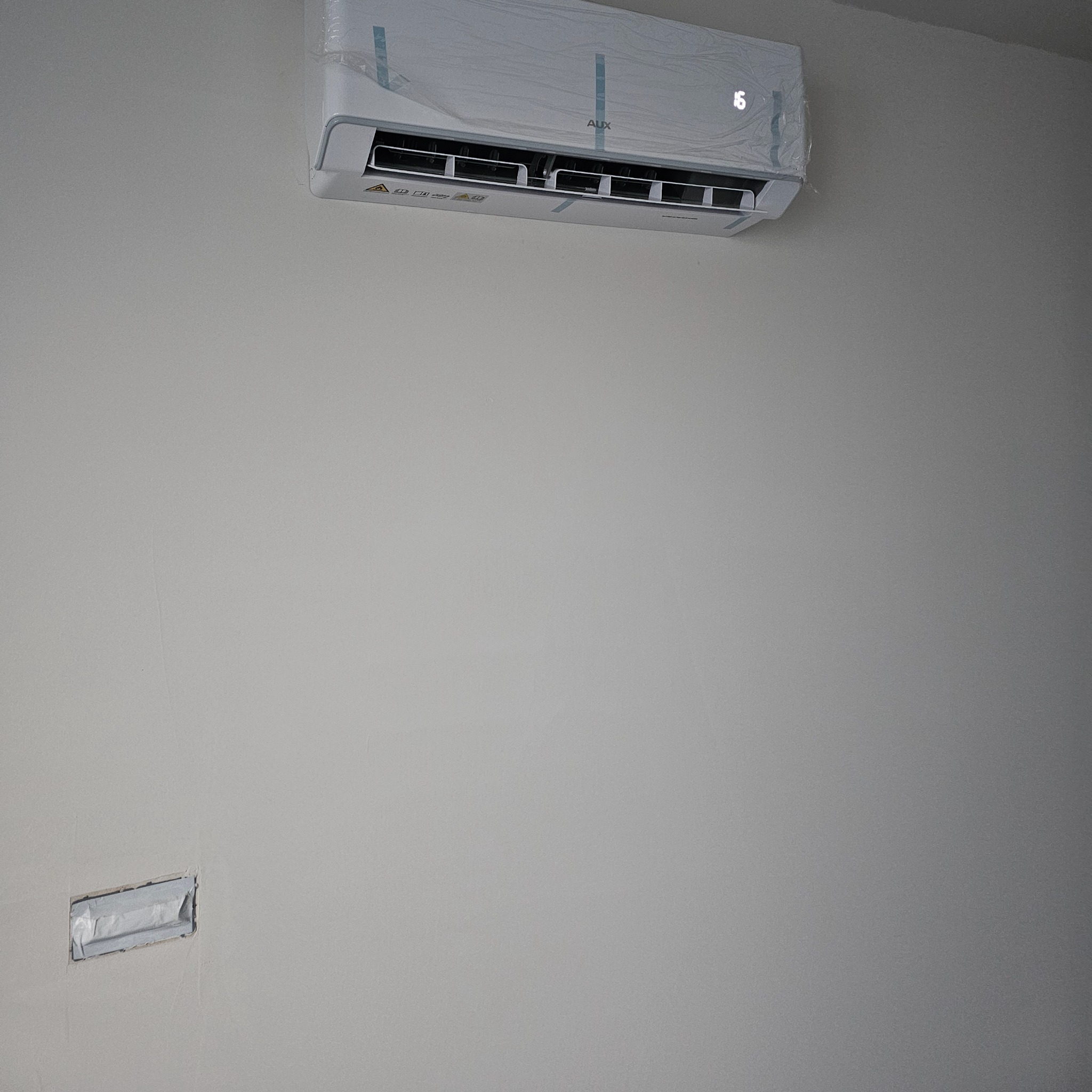 Biały klimatyzator AUX, jeszcze w folii ochronnej, zamontowany na jasnej ścianie. Widoczny fragment przygotowanego otworu na instalację.