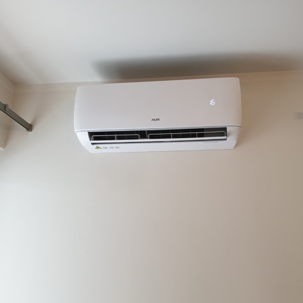 Biała klimatyzacja AUX zamontowana na ścianie o kremowym odcieniu. Widoczny wyświetlacz z temperaturą 16 stopni Celsjusza. Minimalistyczny design urządzenia.