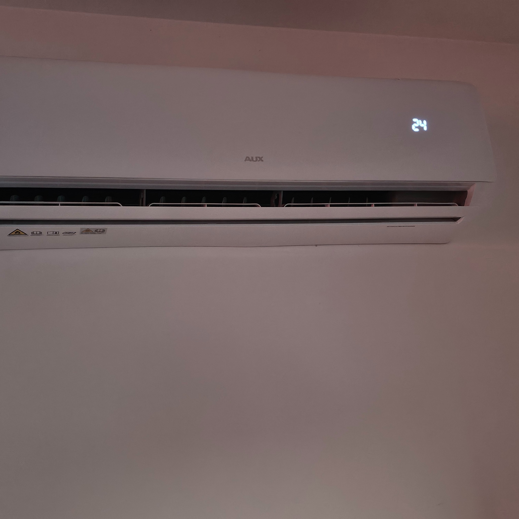 Biała klimatyzacja AUX na ścianie, wyświetlająca temperaturę 24 stopnie. Widoczne detale obudowy i kratki wentylacyjne.
