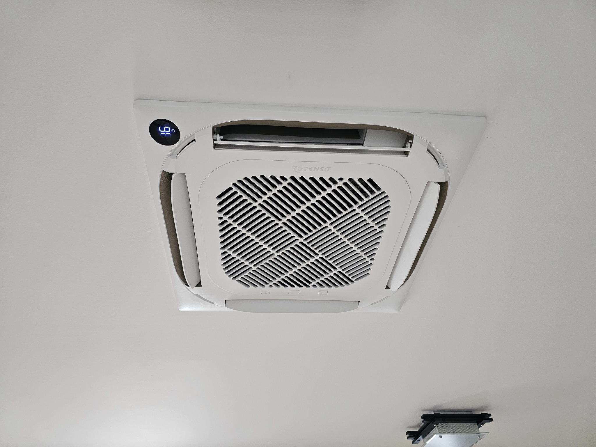 Biała, sufitowa klimatyzacja kasetonowa marki Rotenso z wyświetlaczem temperatury, zamontowana na białym suficie. Widoczny fragment oświetlenia.