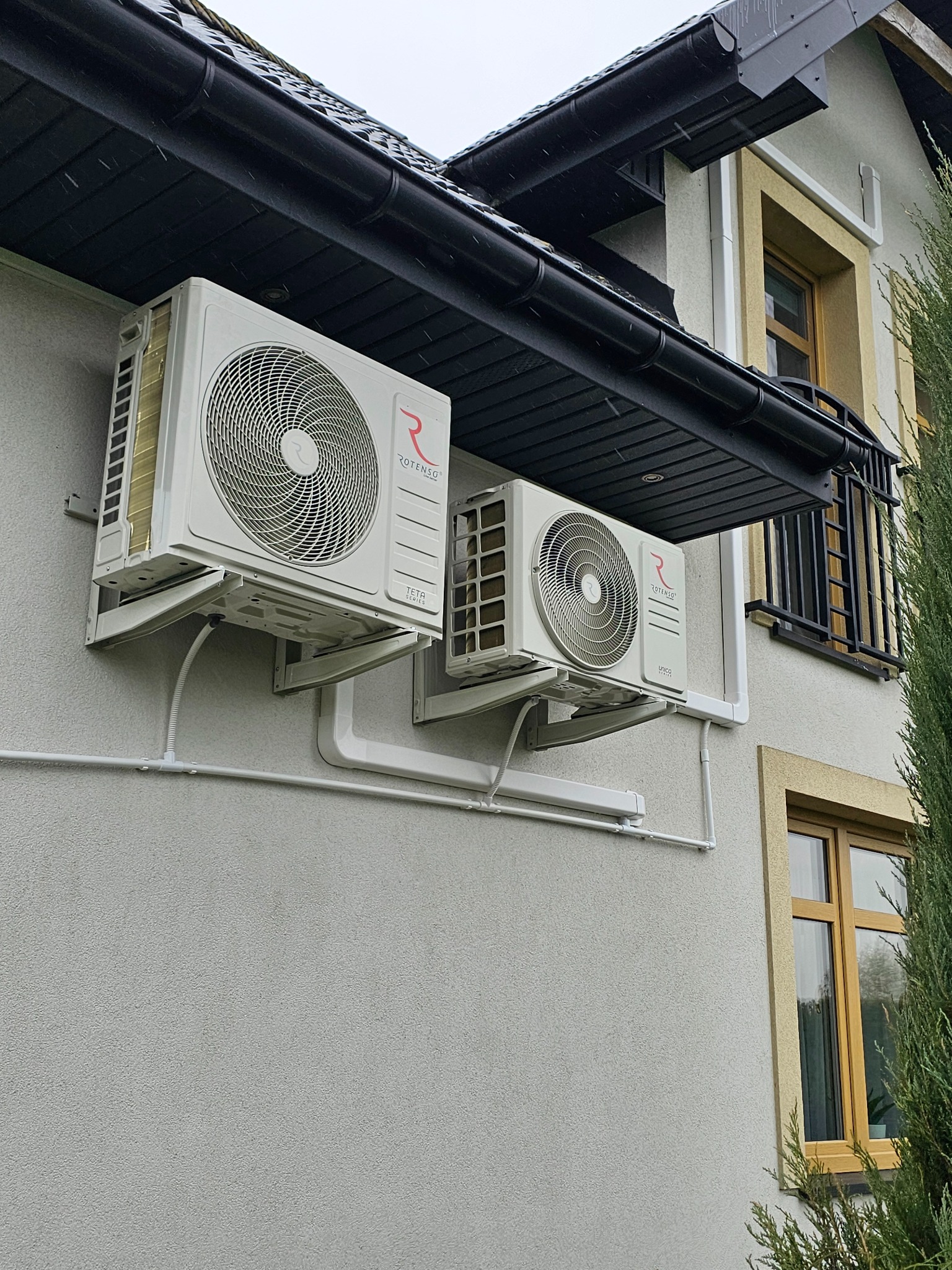 Dwa białe klimatyzatory Rotenso zamontowane na elewacji domu z widoczną instalacją odprowadzającą skropliny. Czarny dach i okno w tle.