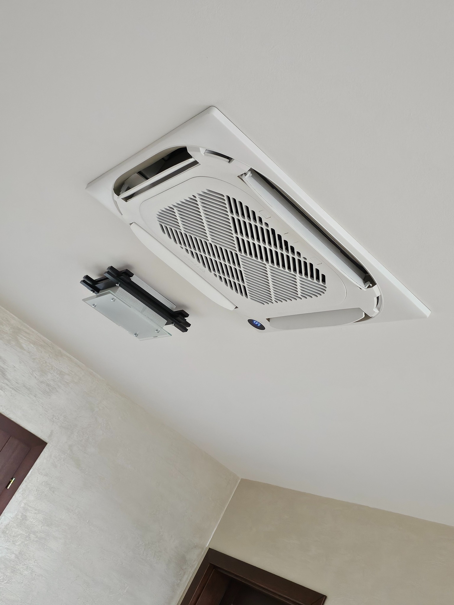 Zamontowany klimatyzator kasetonowy w suficie, z otwartym panelem serwisowym, widok z dołu. Białe urządzenie na tle jasnego sufitu.