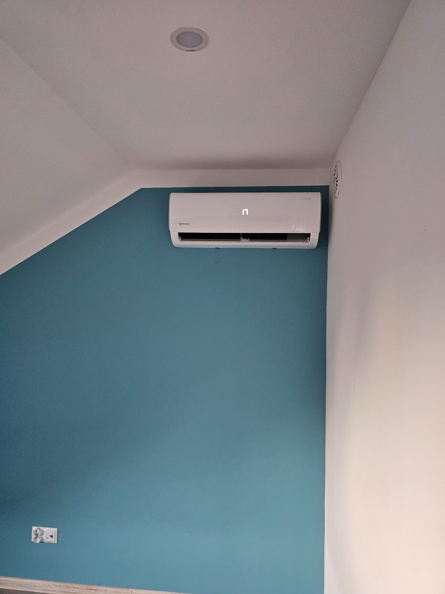 Biała klimatyzacja Rotenso na turkusowej ścianie w pokoju na poddaszu. Widoczny fragment gniazdka elektrycznego i kratka wentylacyjna.