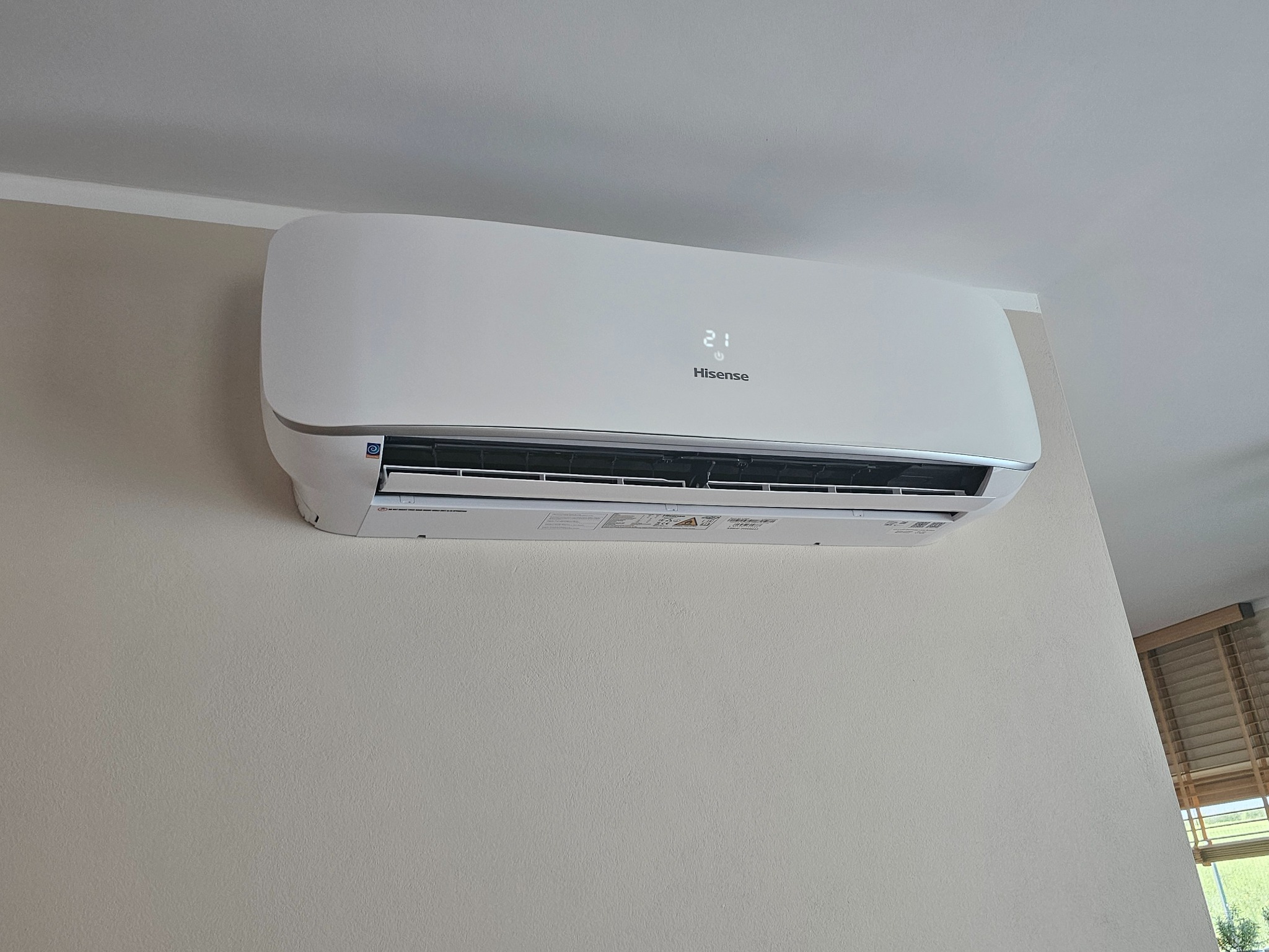 Biały klimatyzator Hisense z wyświetlaną temperaturą 21°C, zamontowany na jasnej ścianie w pomieszczeniu z żaluzjami w tle.