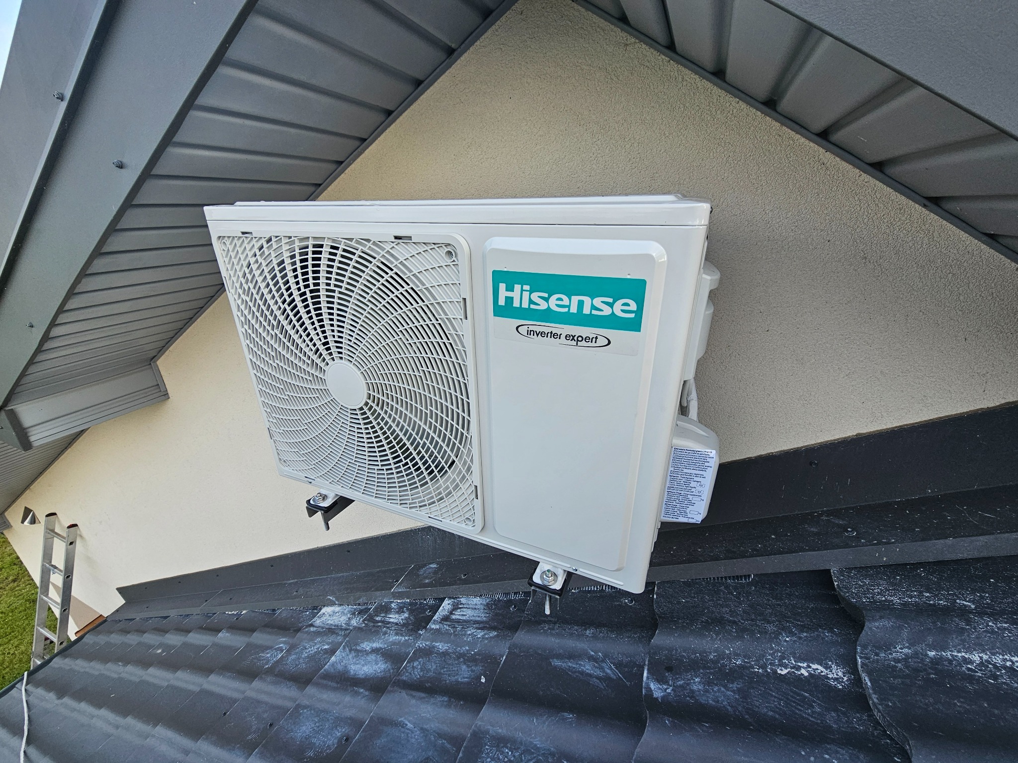 Biała jednostka zewnętrzna klimatyzacji Hisense zamontowana na elewacji domu, widoczna z dachu. Obok drabina. Ujęcie z dołu.
