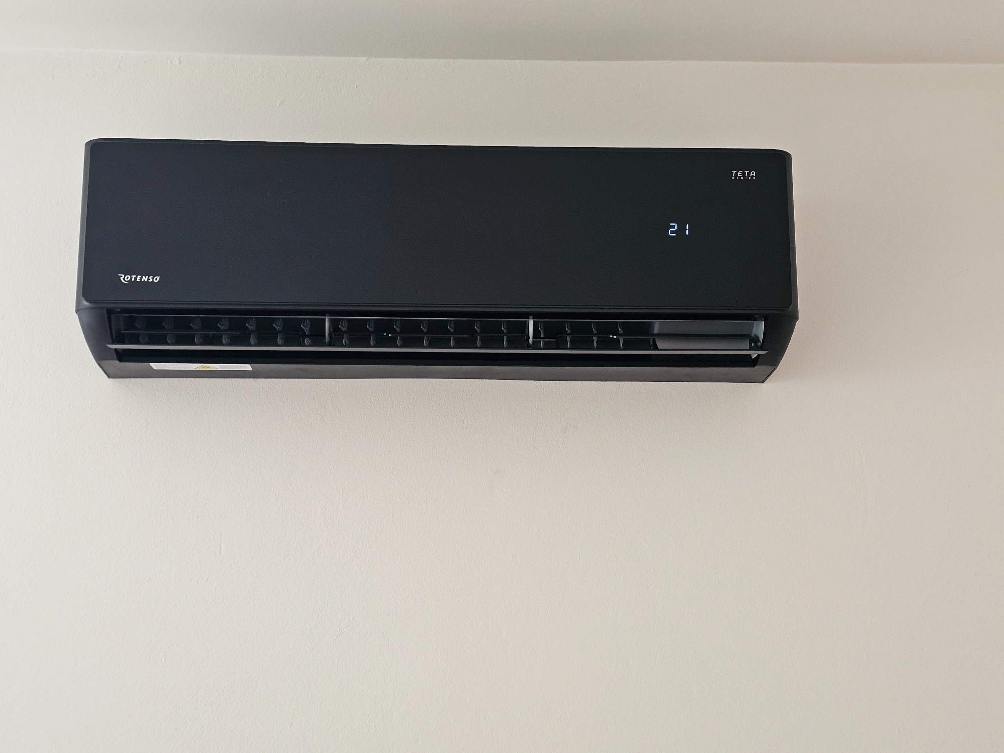 Czarny klimatyzator Rotenso Teta na kremowej ścianie. Wyświetlacz pokazuje 21 stopni. Minimalistyczny design, nowoczesne wnętrze.