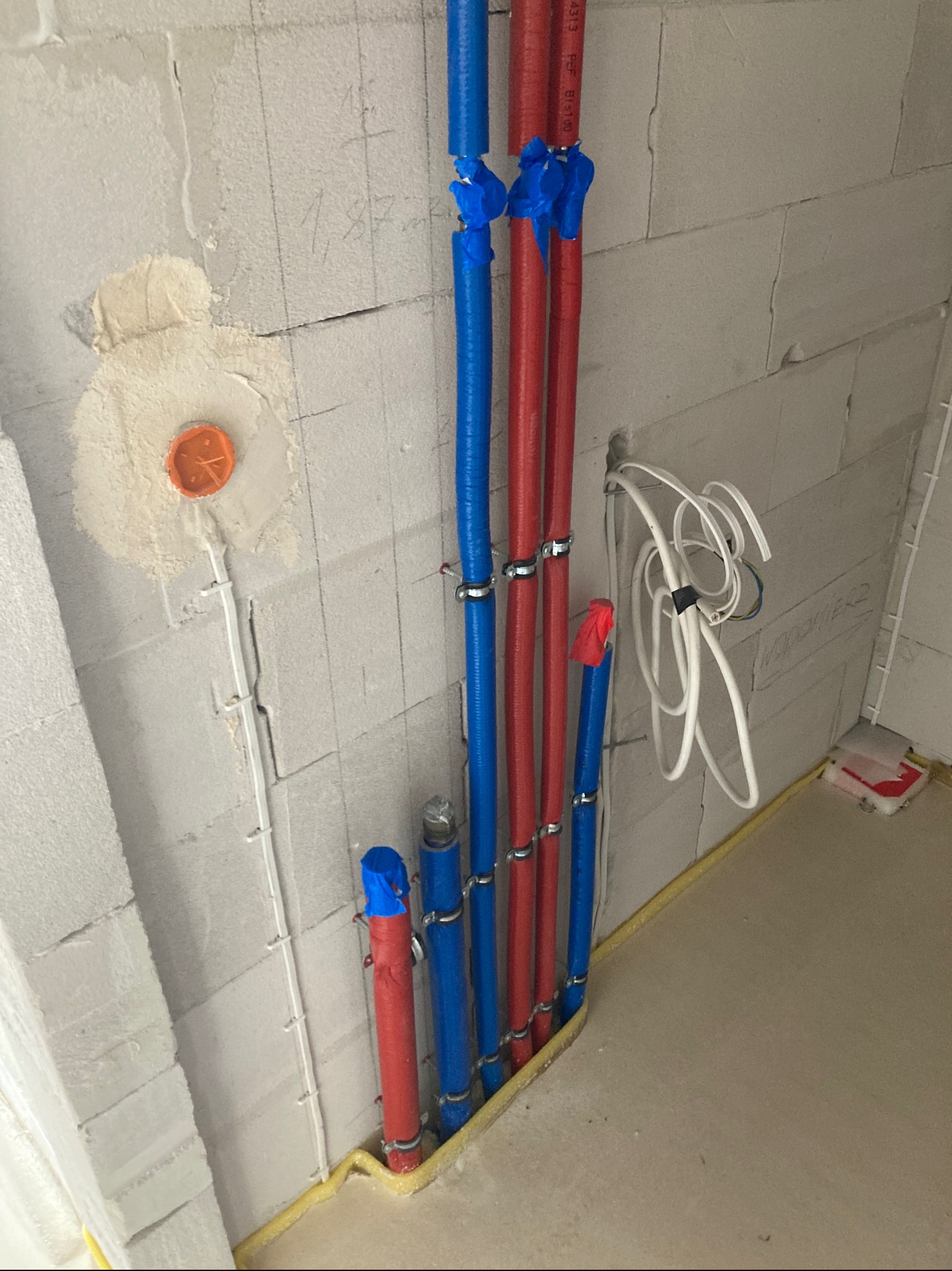 Instalacja wodna w budynku w trakcie budowy: rury czerwone i niebieskie, zabezpieczone taśmą, przymocowane do ściany z betonu komórkowego. Widoczne przewody elektryczne.