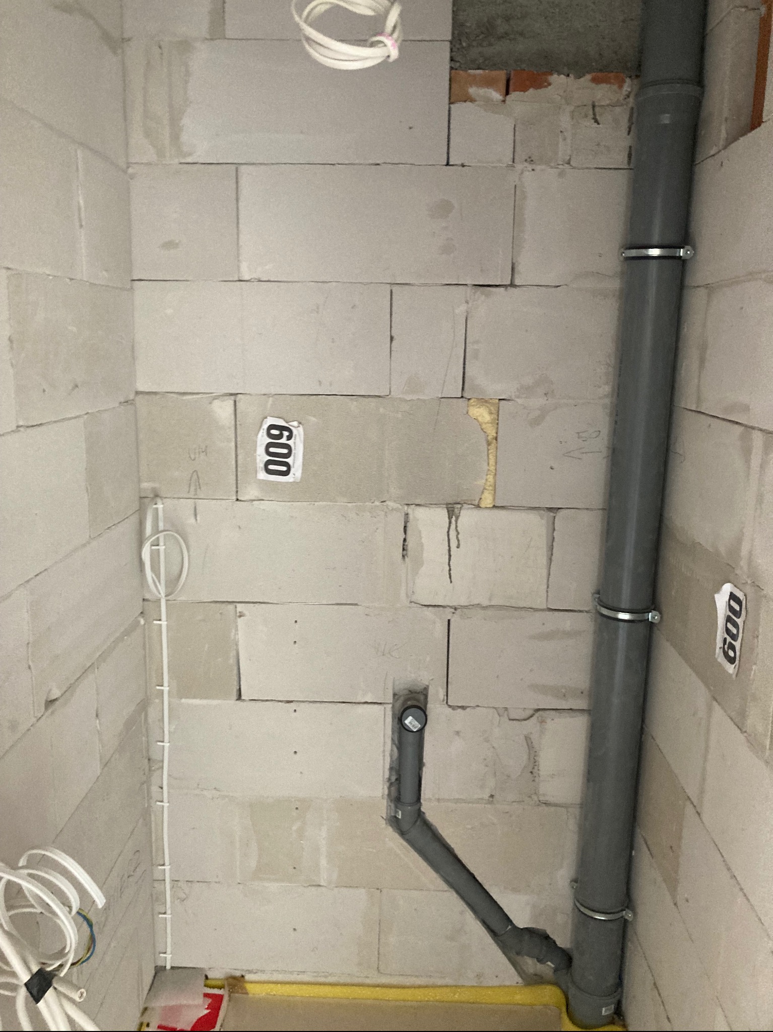 Szare rury kanalizacyjne przymocowane do ściany z betonu komórkowego. Widoczne także przewody elektryczne i fragment żółtej rury w podłodze. Stan surowy.
