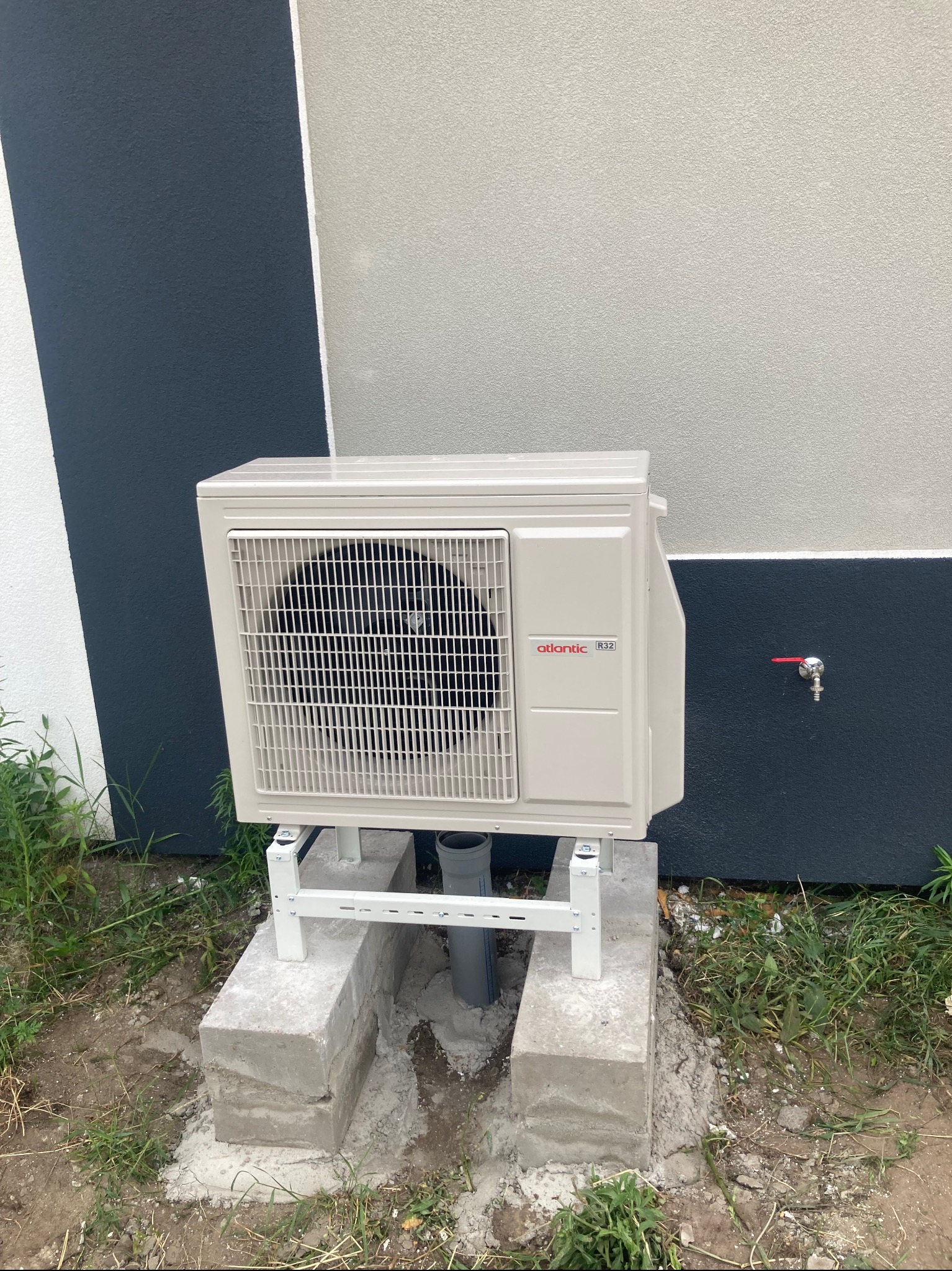 Beżowa zewnętrzna jednostka klimatyzacji Atlantic R32, zamontowana na betonowym fundamencie przy ścianie budynku z kranem. Widoczne trawy i ziemia.