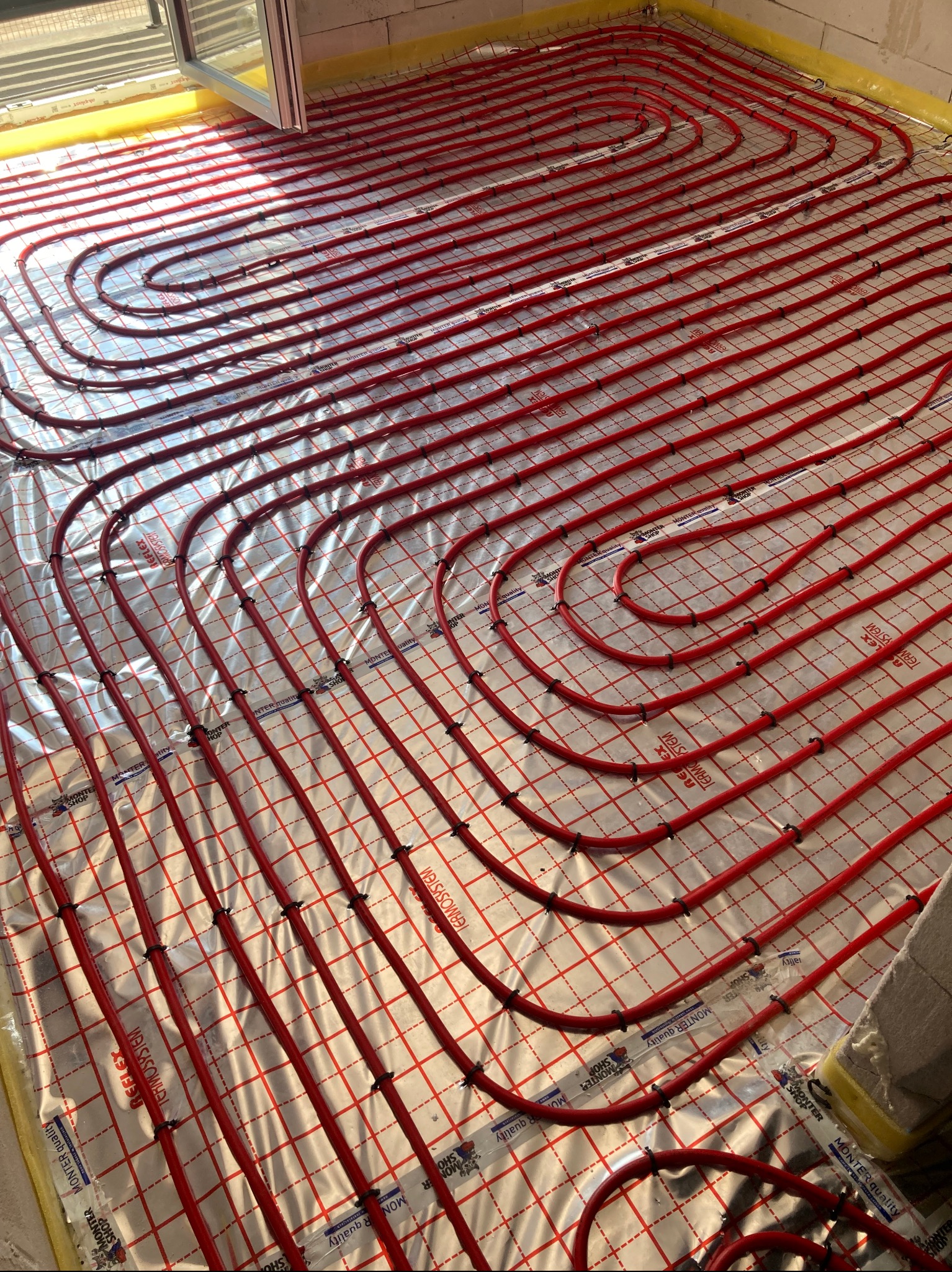 Czerwone rury ogrzewania podłogowego ułożone na srebrnej folii z nadrukiem, widok z góry, w tle okno i ściana z betonu komórkowego.