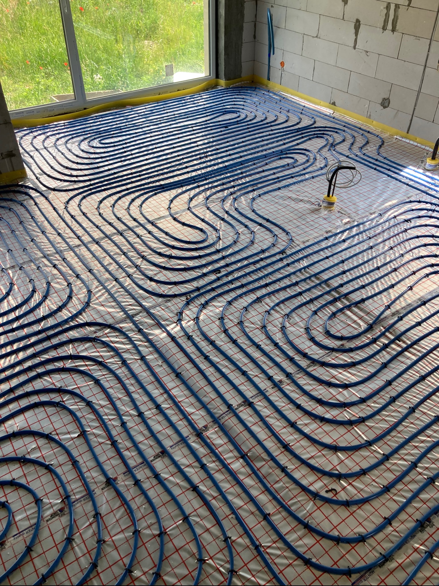 Niebieskie rury ogrzewania podłogowego ułożone w pętle na folii z siatką w surowym wnętrzu. Widoczne okno i fragment ściany z bloczków. Żółta taśma izolacyjna.
