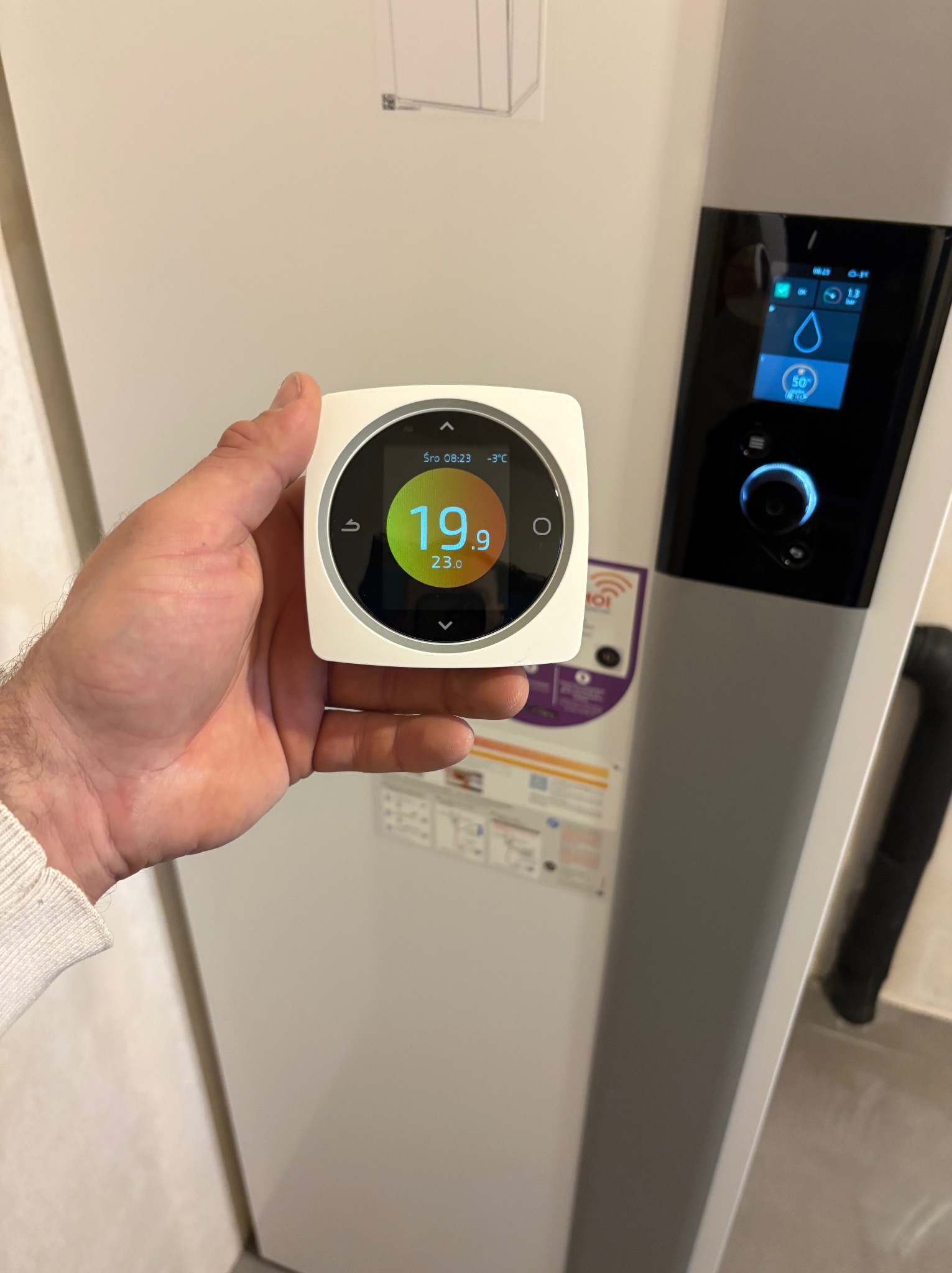 Biały, dotykowy termostat w dłoni na tle pieca gazowego z wyświetlaczem. Widoczna temperatura 19.9°C. Nowoczesny system sterowania ogrzewaniem.