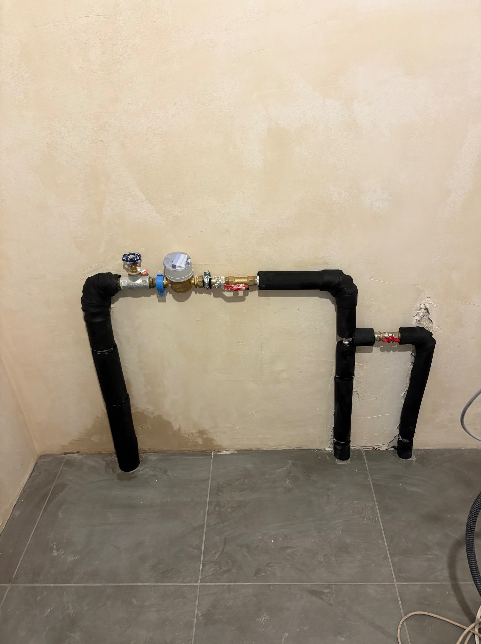 Kompleksowa instalacja wodna z licznikiem, rury zaizolowane czarną otuliną, szare płytki podłogowe, beżowa ściana z widocznymi śladami po pracach instalacyjnych.