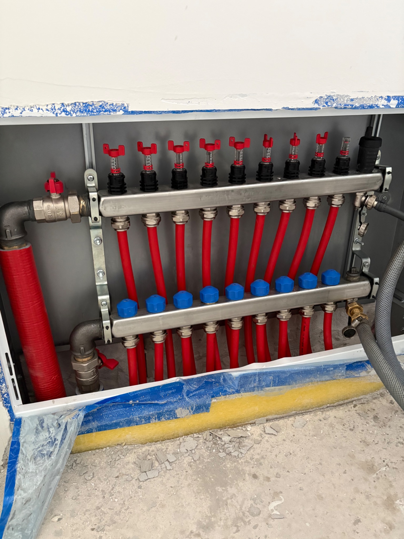 Rozdzielacz ogrzewania podłogowego z czerwonymi rurami i niebieskimi zaworami, zamontowany w szarej szafce, widoczne zabezpieczenia podłoża.