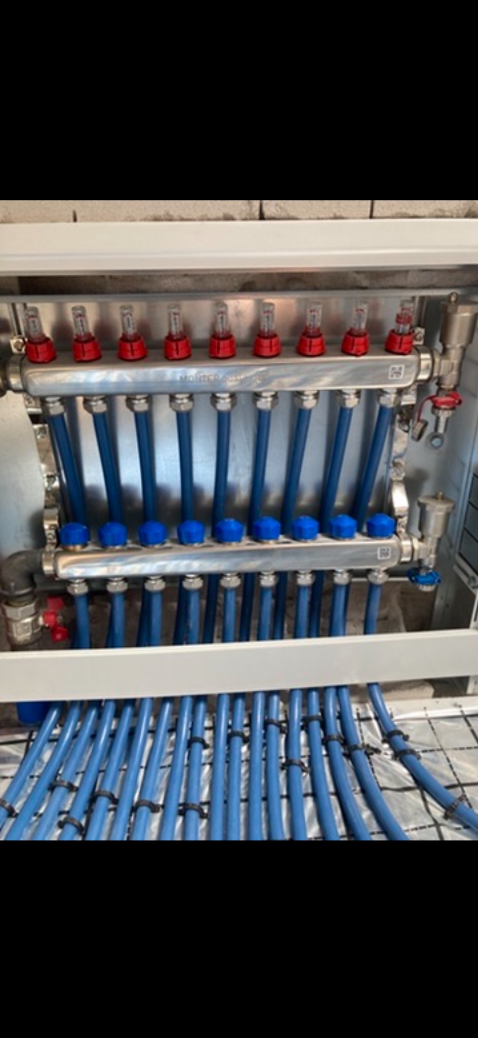 Rozdzielacz ogrzewania podłogowego z niebieskimi rurami, zamontowany w szafce, widoczne elementy regulacyjne i mocowania rur do maty izolacyjnej.