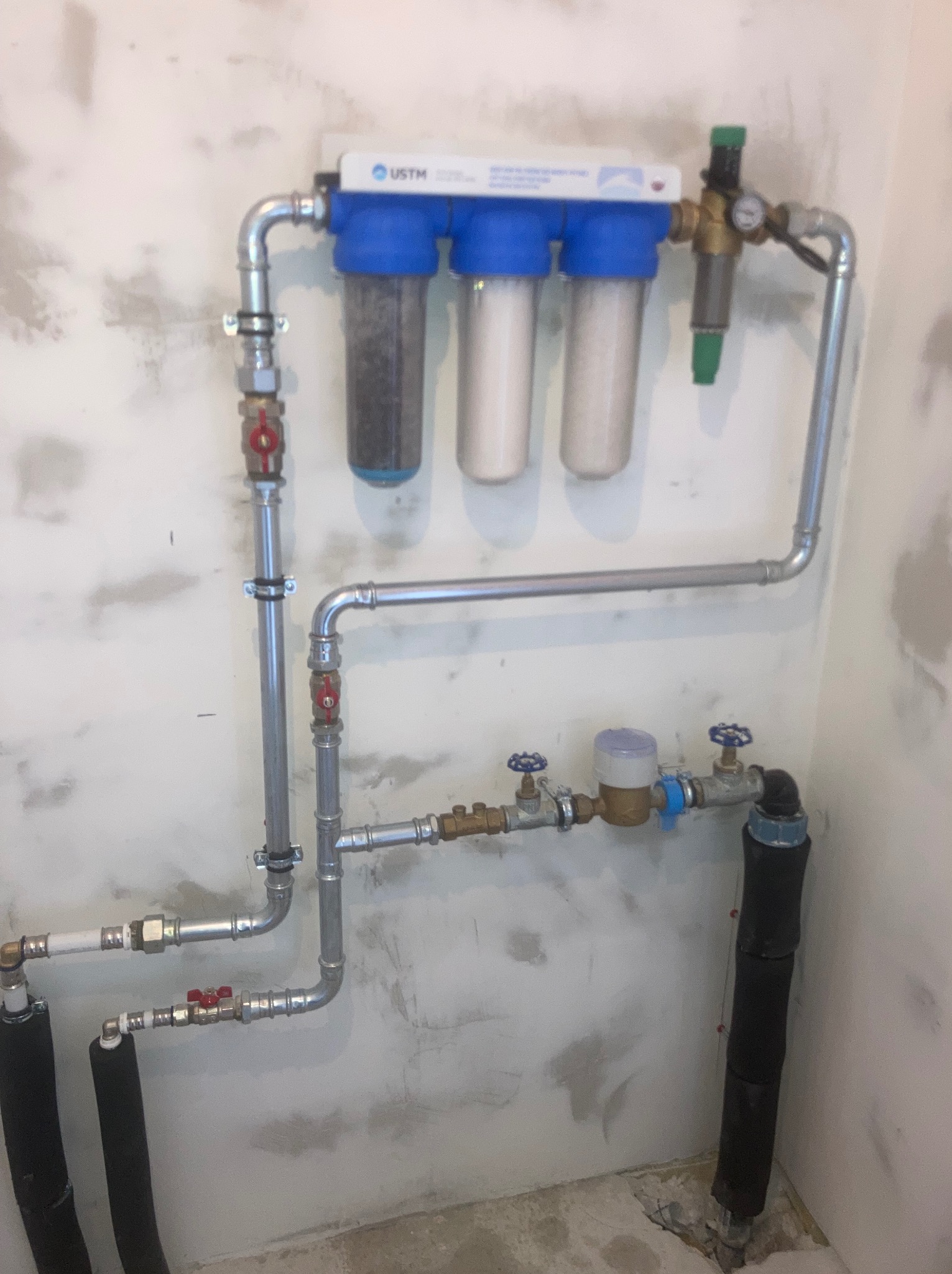 Zamontowany system filtrów wody z licznikiem, rurami i zaworami na tle surowej ściany. Widoczne elementy instalacji hydraulicznej.