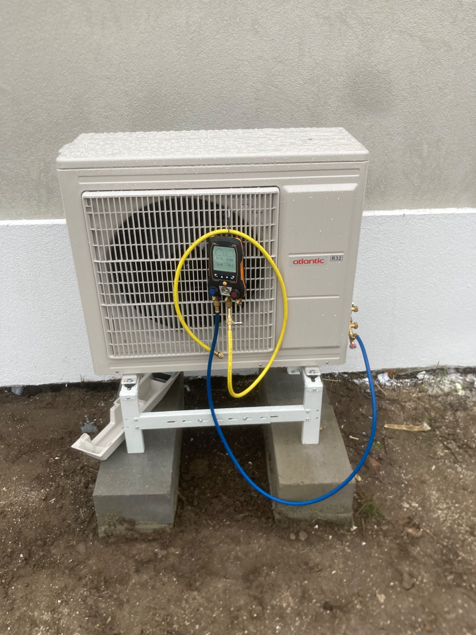 Klimatyzator Atlantic R32 na betonowych podstawach, podłączony do manometru z żółtymi i niebieskimi przewodami, na tle szarej ściany w deszczowy dzień.