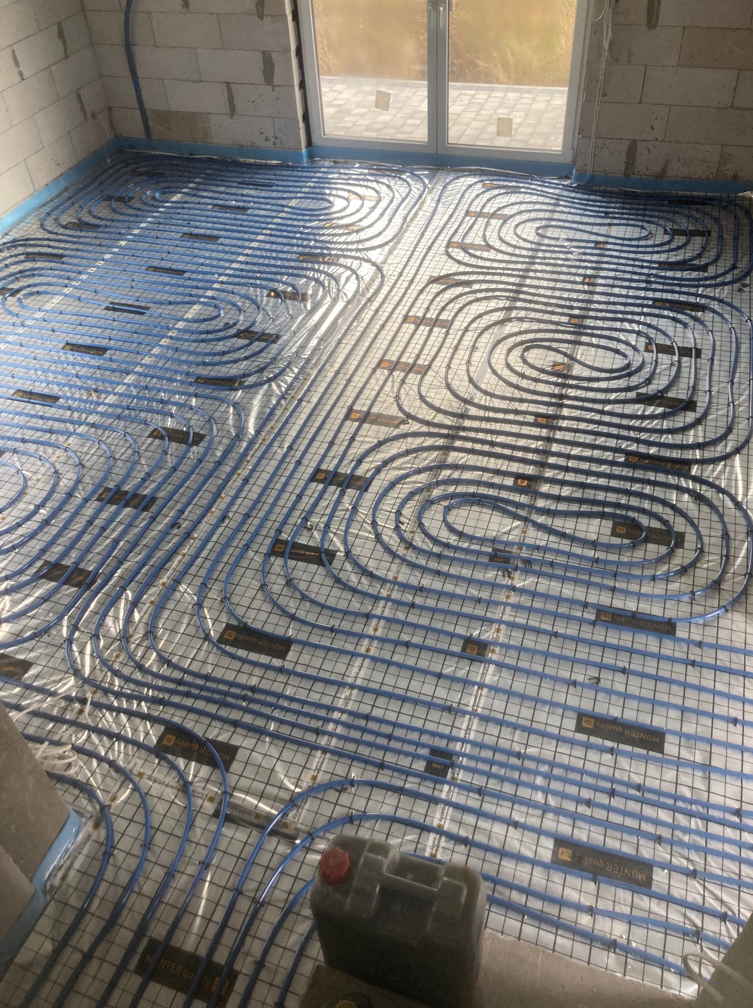 Niebieskie rury ogrzewania podłogowego ułożone na folii w surowym wnętrzu, widoczne elementy mocujące i fragment ściany z bloczków. Etap instalacji przed wylewką.