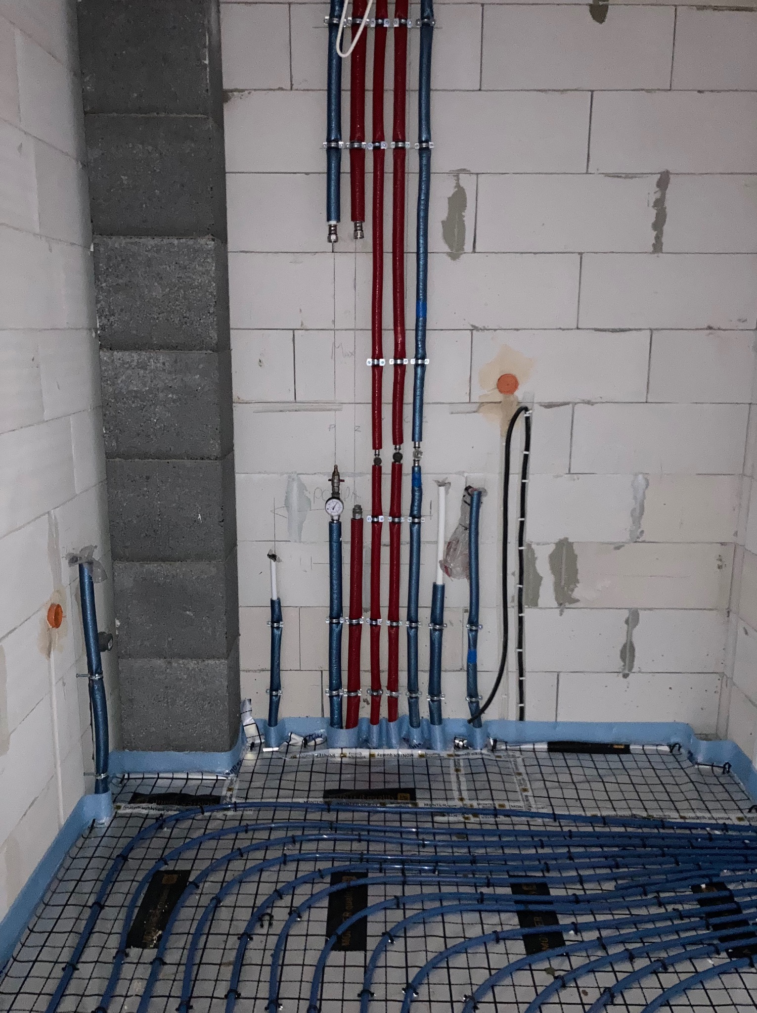 Niebieskie rury ogrzewania podłogowego ułożone na siatce w surowym pomieszczeniu z białymi ścianami z betonu komórkowego i pionowymi rurami w kolorze czerwonym i niebieskim.