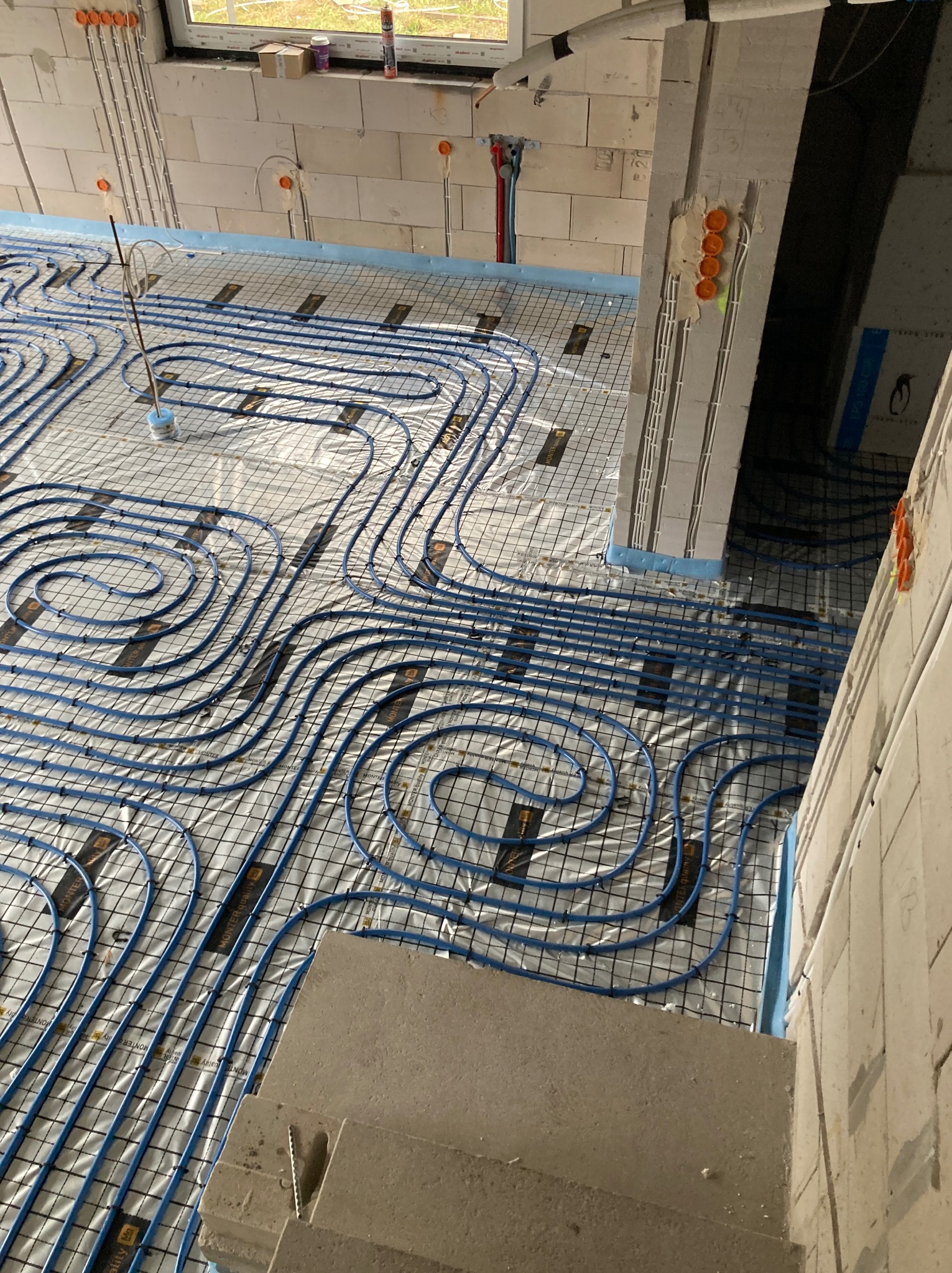 Niebieskie rury ogrzewania podłogowego ułożone na siatce w surowym wnętrzu, widoczne przewody elektryczne i okno w tle. Instalacja przed wylaniem posadzki.